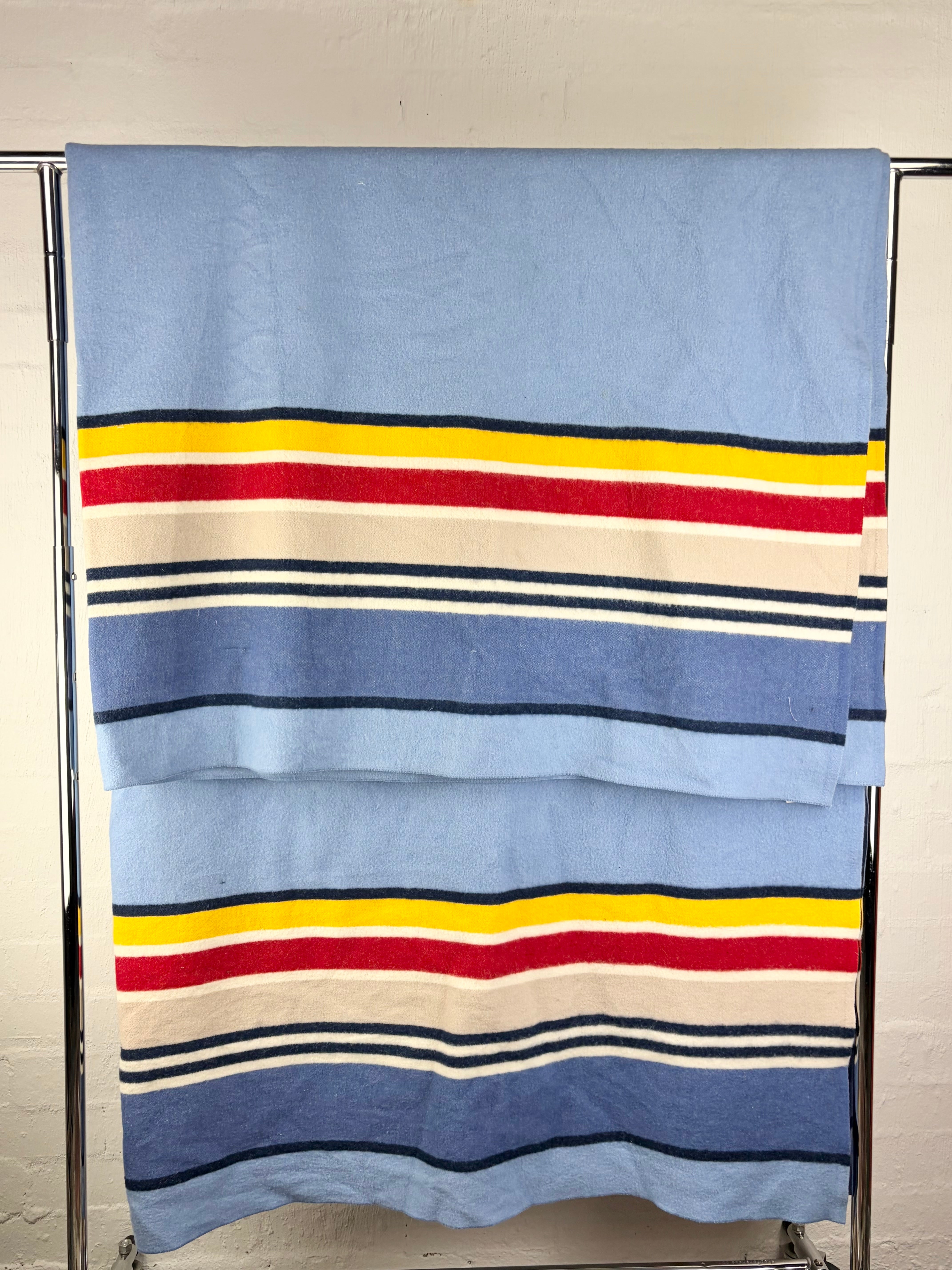 A folded blue striped textile, likely a towel or blanket, hanging on a metal rack against a white wall. The fabric features horizontal stripes in navy, cream, red, and yellow.