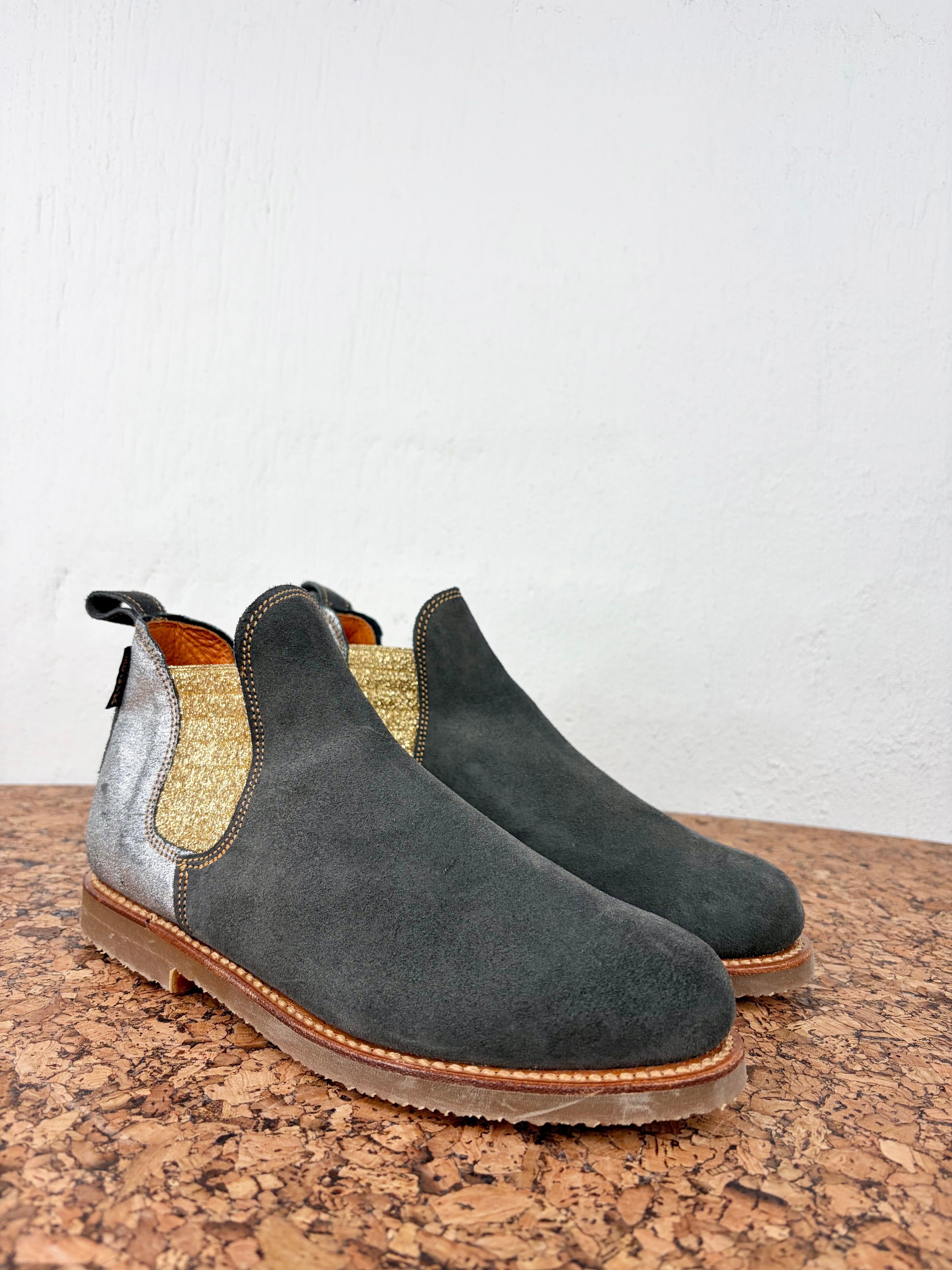 A pair of dark gray suede Chelsea boots with metallic silver side panels and a textured gold elastic side gore, resting on a cork surface against a white wall.
