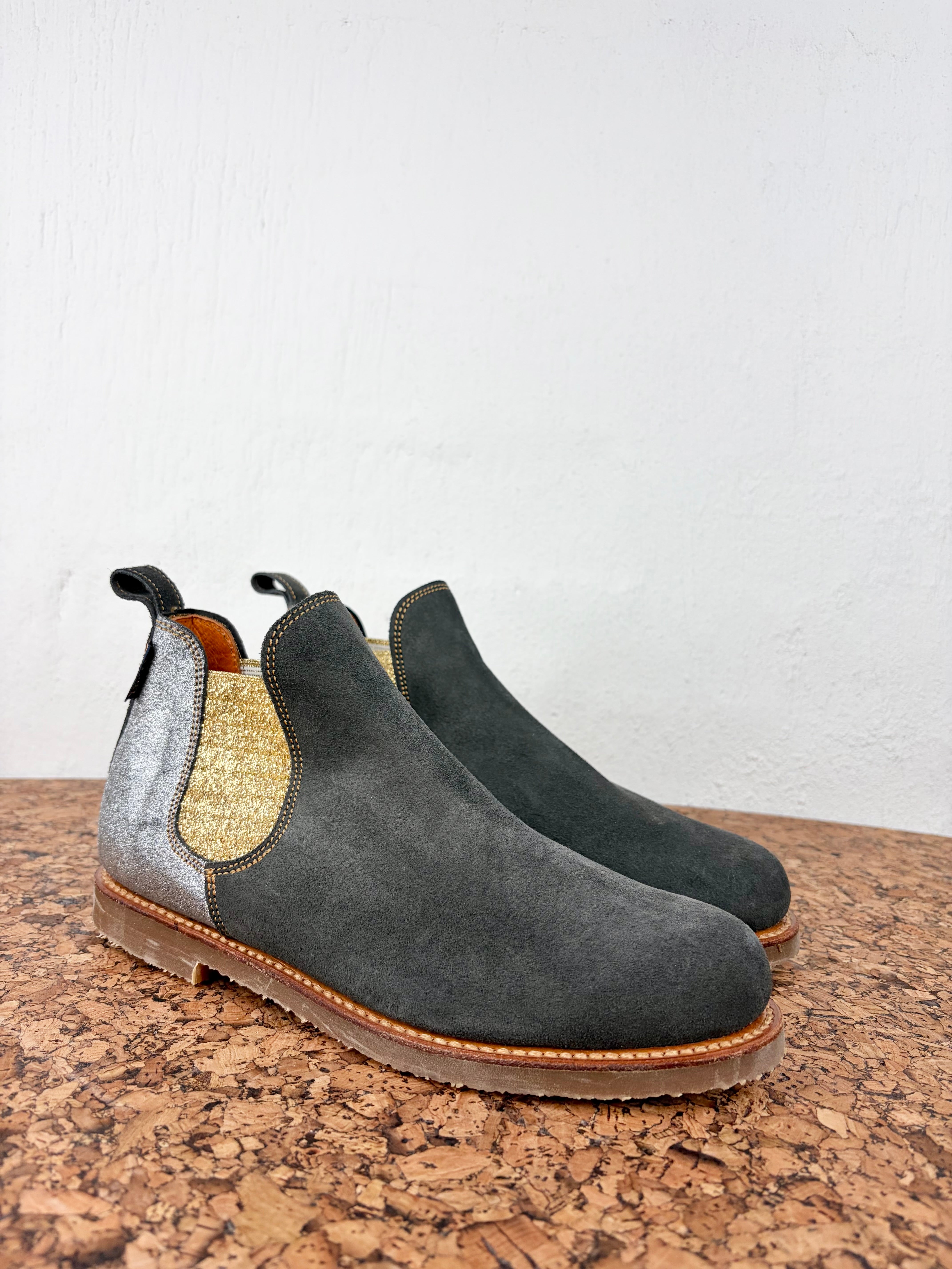 A pair of Chelsea-style ankle boots in dark gray suede with a textured gold side panel and elastic side gore, resting on a cork surface against a white wall.
