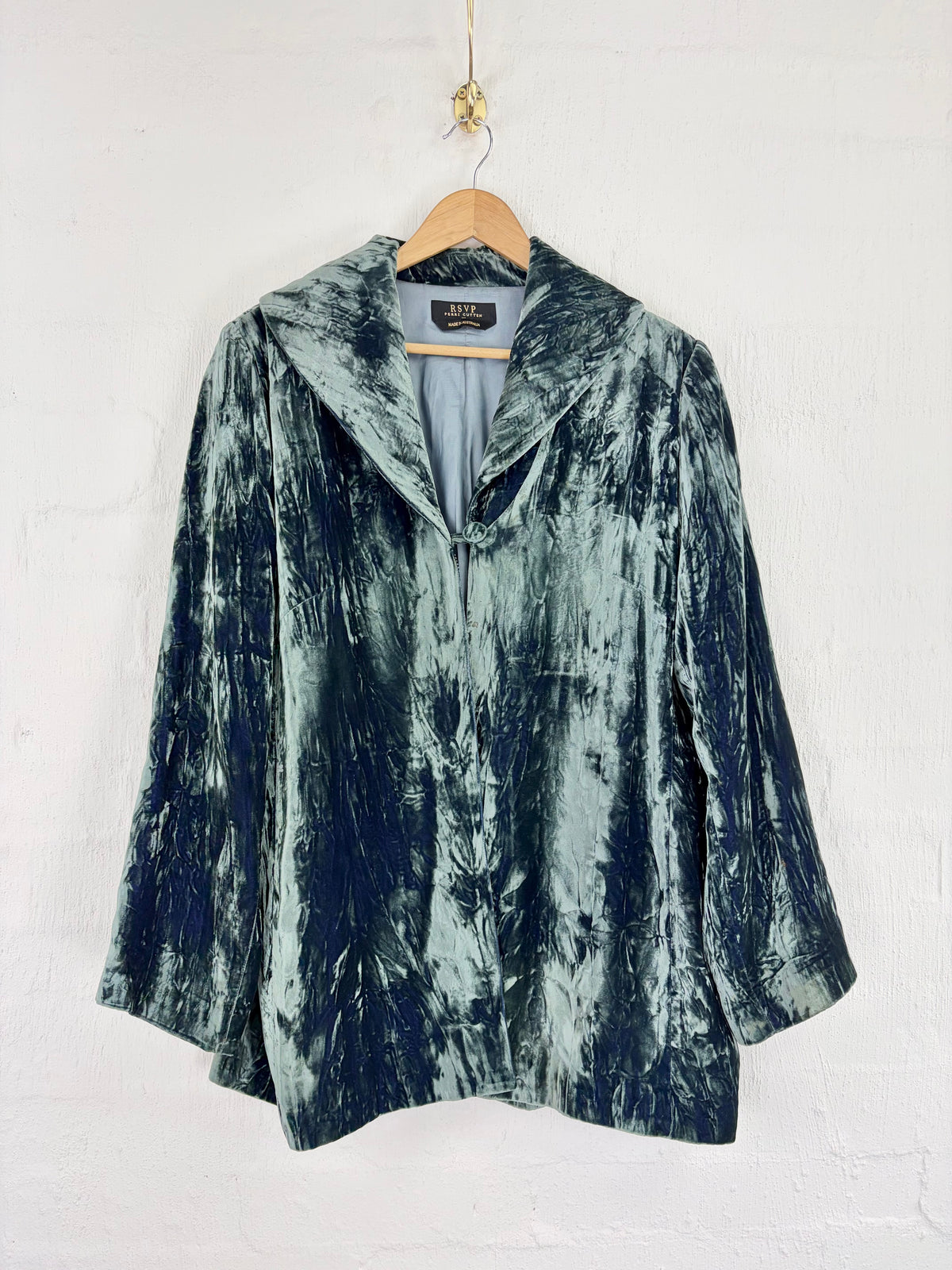 A dark teal and navy blue marbled velvet jacket with a wide collar, hanging on a wooden hanger against a white painted brick wall.