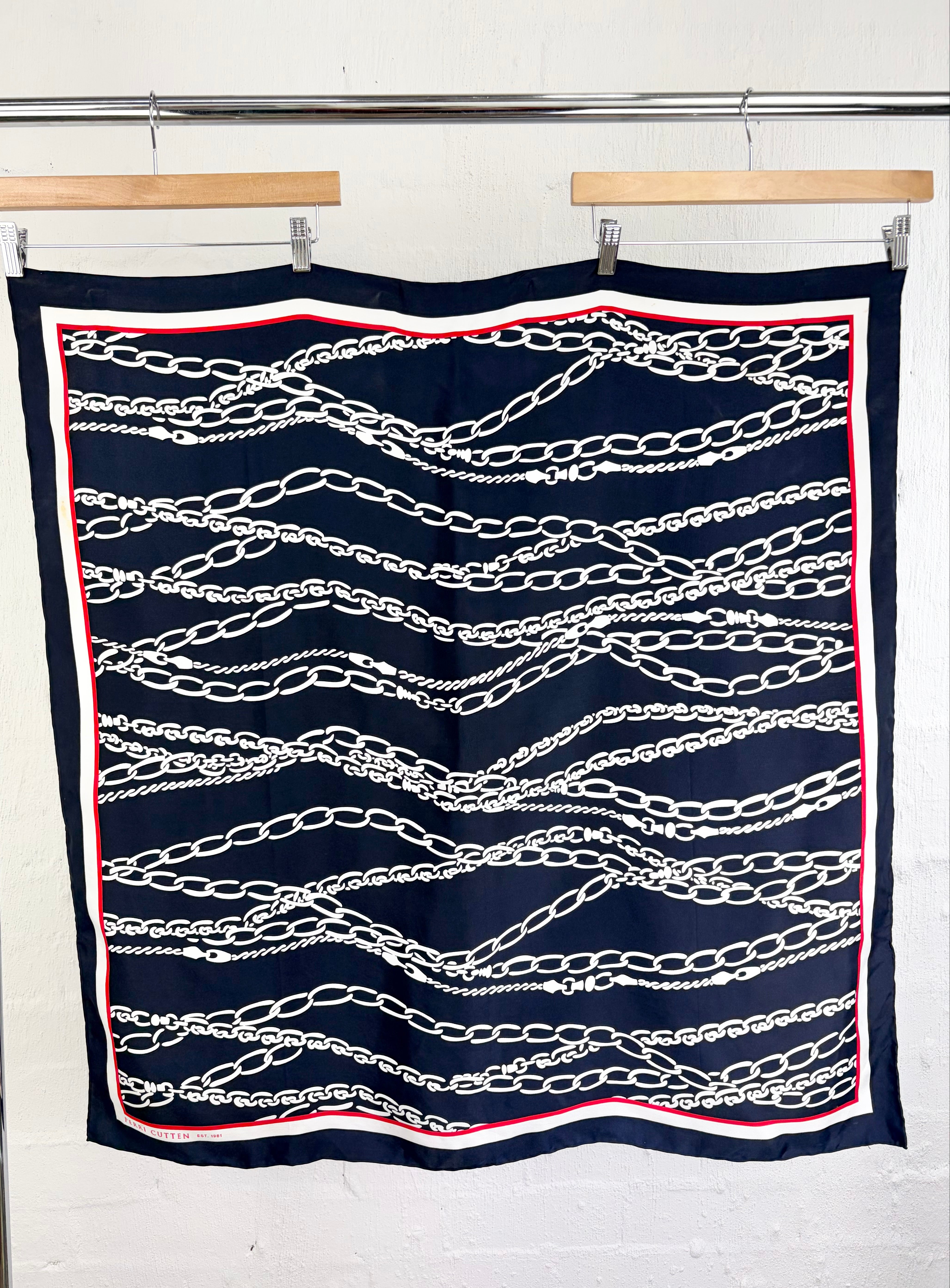 A square navy blue scarf with a white chain-link pattern and a red and white border, hanging from two wooden hangers on a metal rack against a white brick wall.
