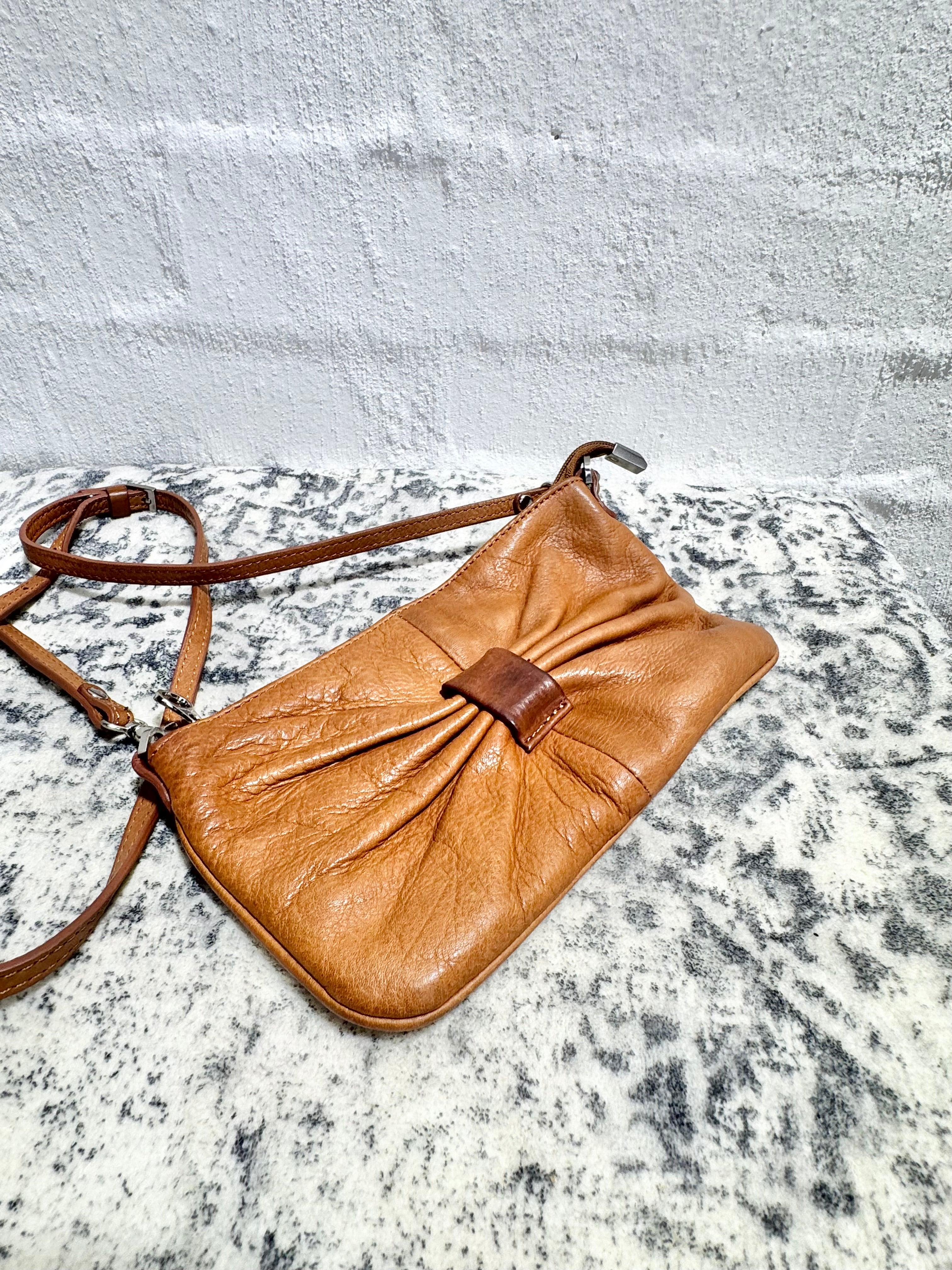 A tan leather crossbody bag with a gathered front and a matching shoulder strap, resting on a gray and white speckled surface against a white brick wall.