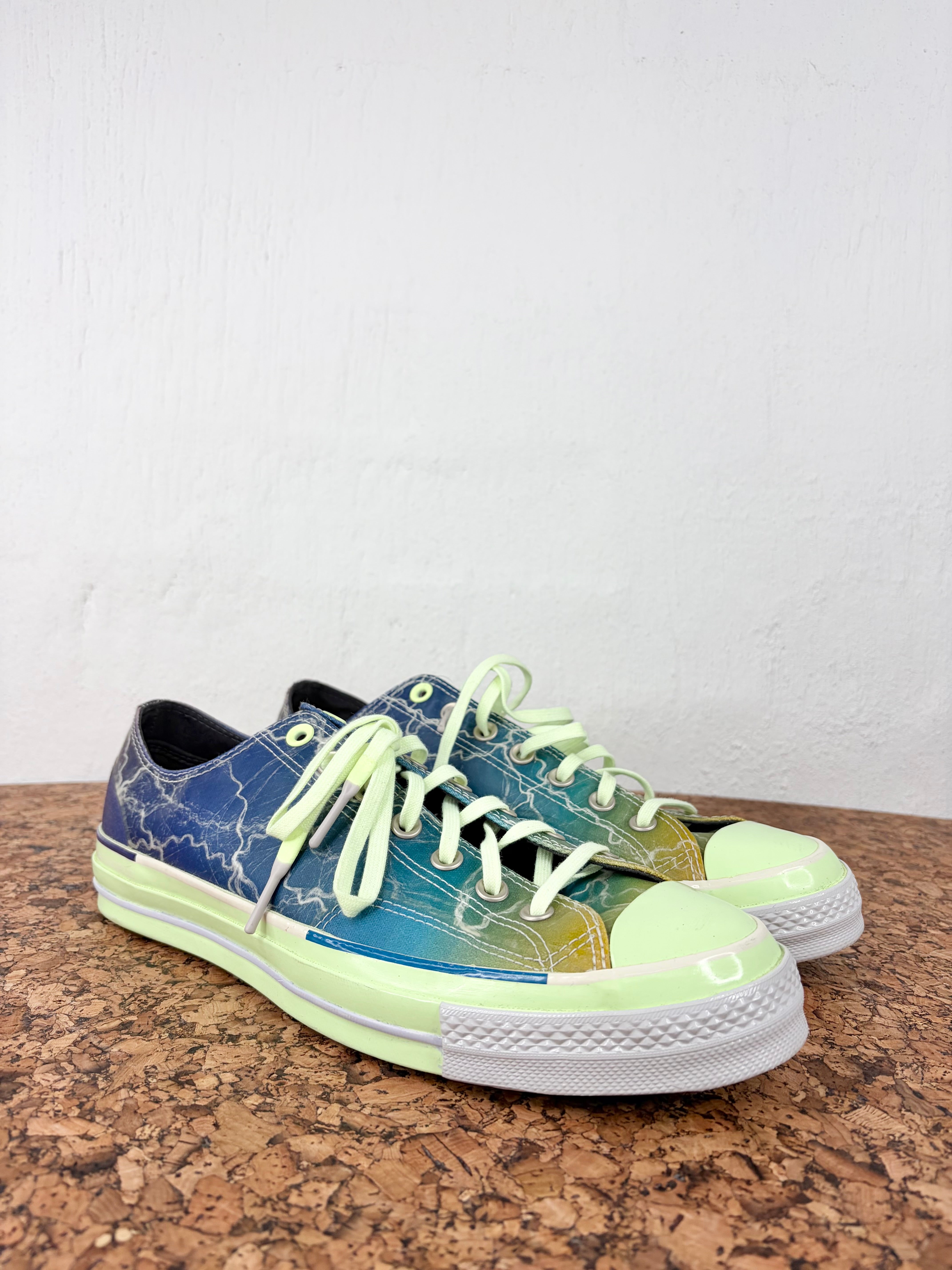 A pair of low-top canvas sneakers with a blue-to-yellow gradient and lightning-bolt pattern, featuring lime green laces, a lime green toe cap, and a white rubber sole, displayed on a cork surface against a white wall.