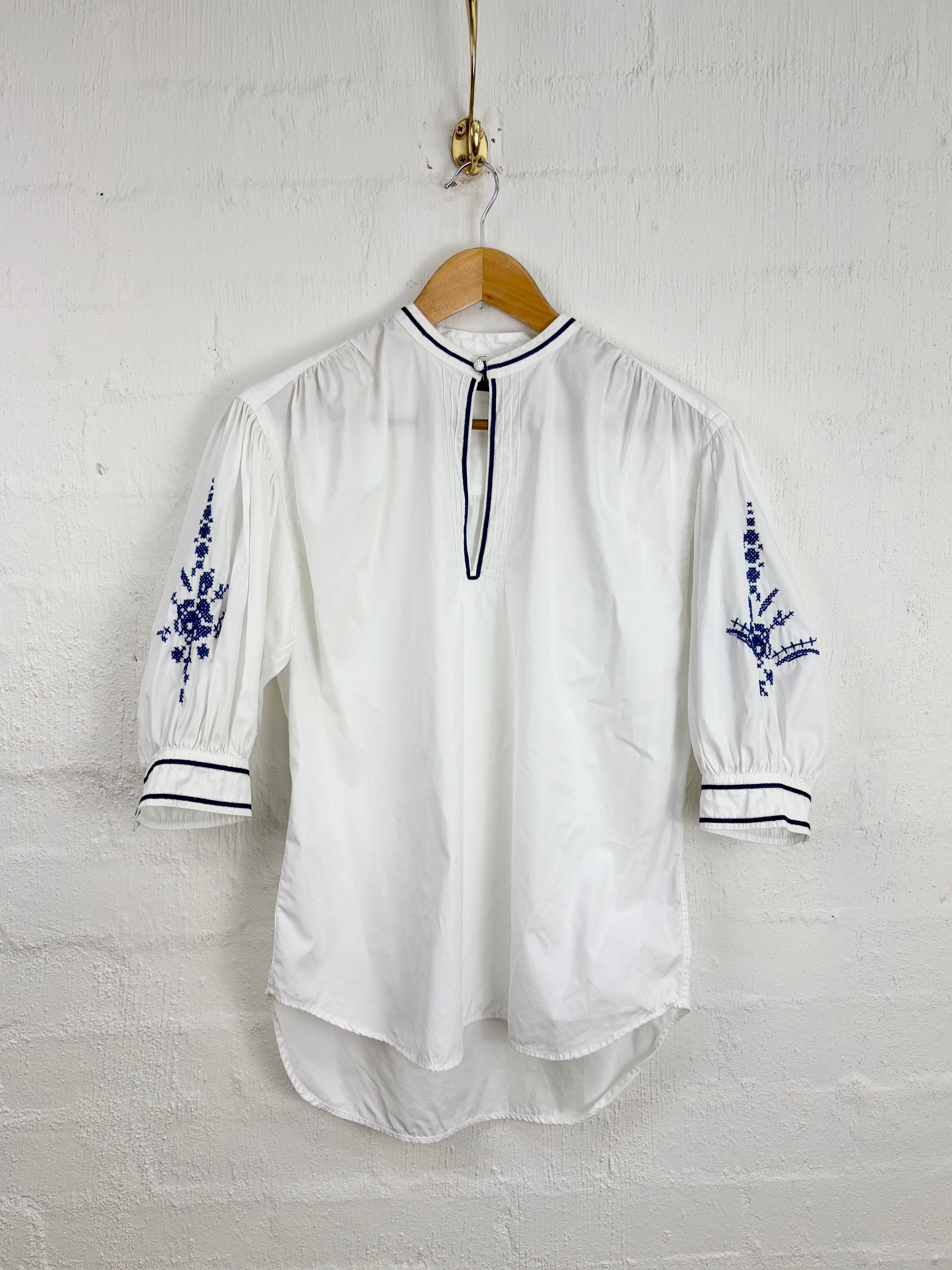 A white blouse with blue floral embroidery on the sleeves and thin black piping details, hanging on a wooden hanger against a white brick wall.