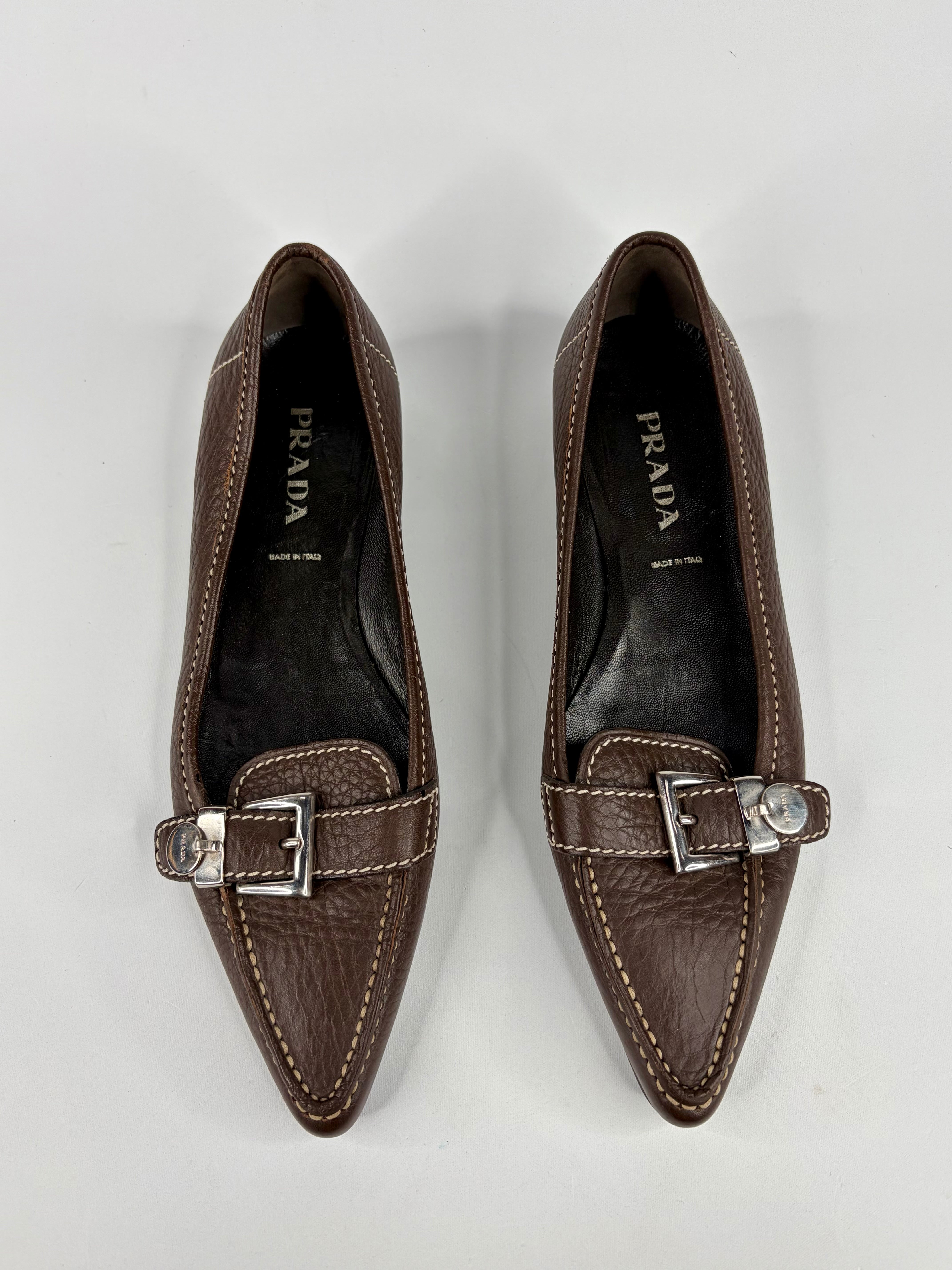 Pair of brown Prada loafers with silver buckles on a light gray background