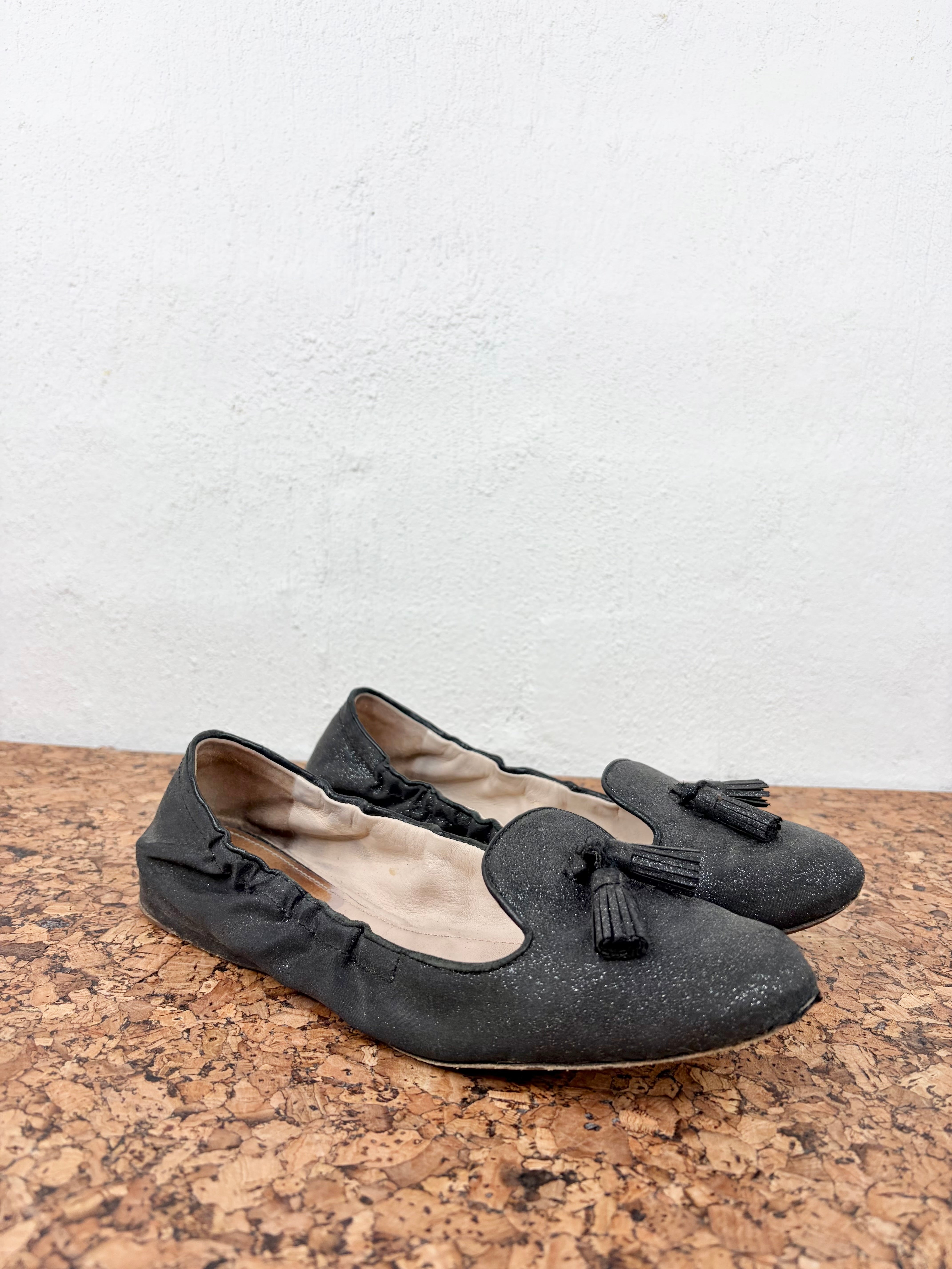 A pair of black flat shoes with ruffled trim and small tassels on the toes, resting on a cork surface against a white wall.
