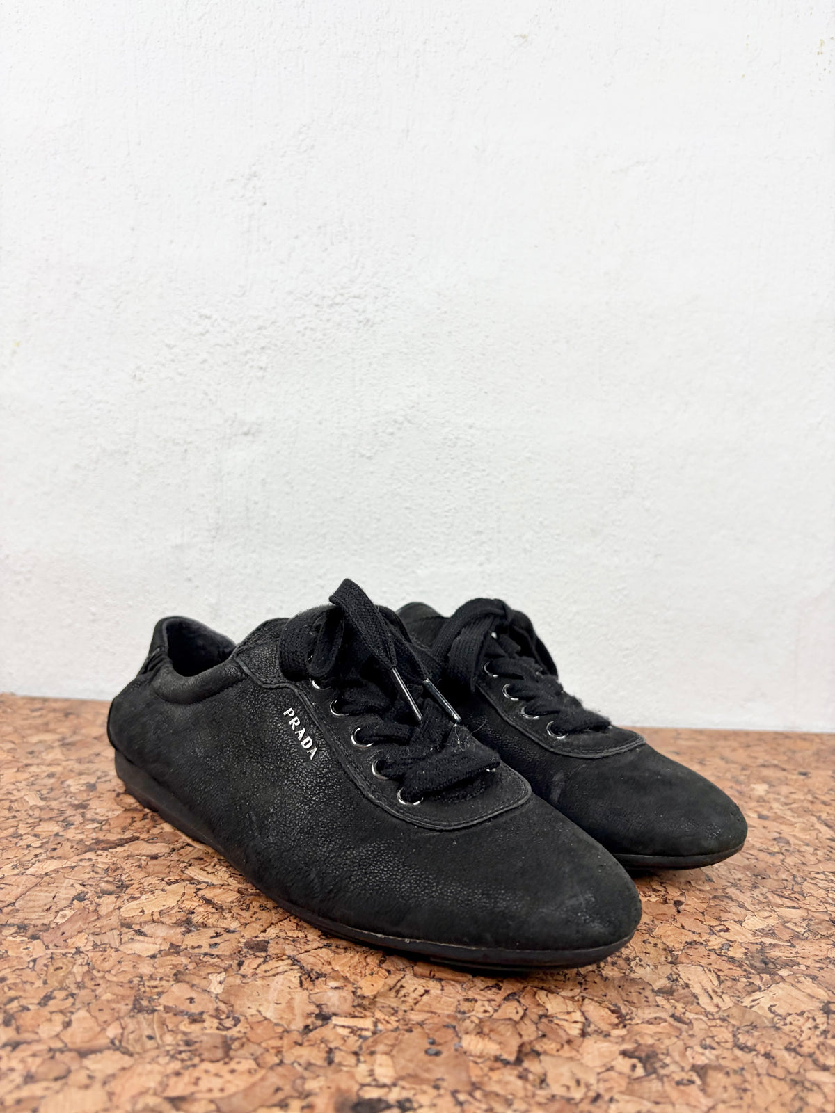 A pair of black suede Prada sneakers with black laces, resting on a cork surface against a white textured wall.