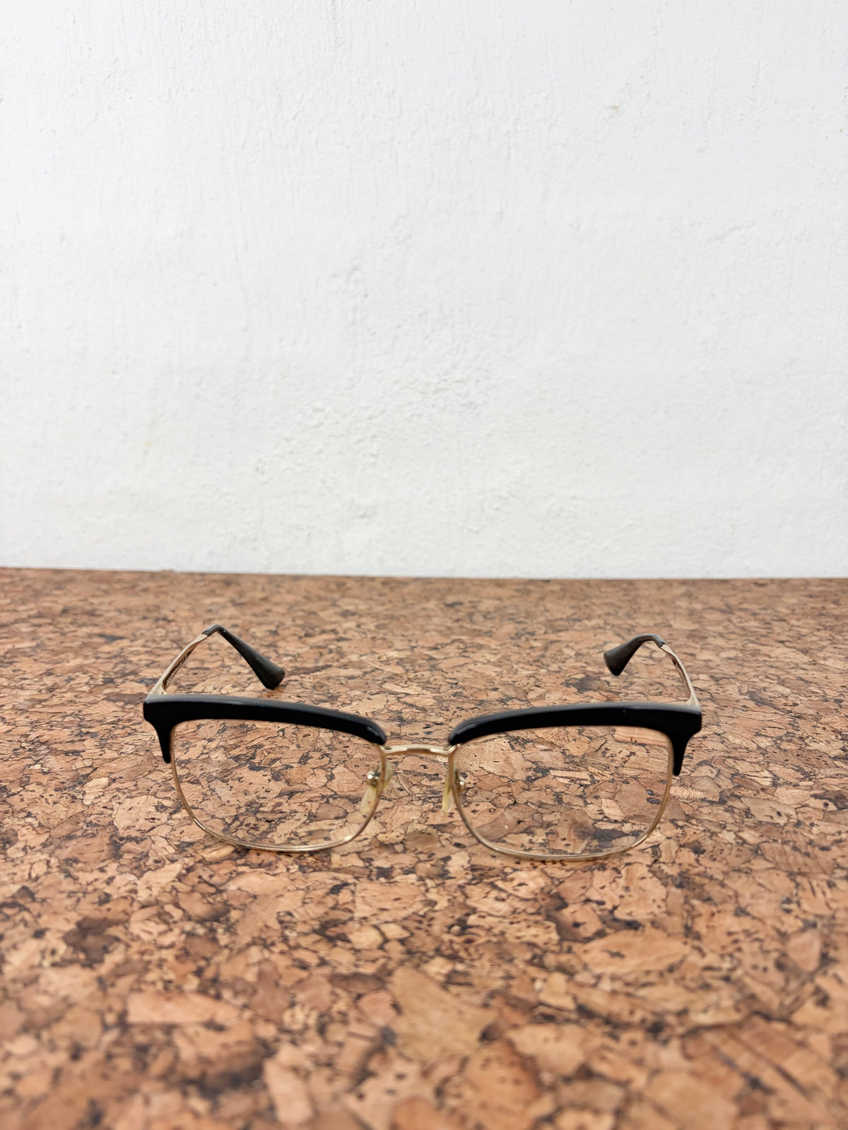 A pair of semi-rimless eyeglasses with black acetate front frames and gold metal temples, resting on a cork surface against a white wall.