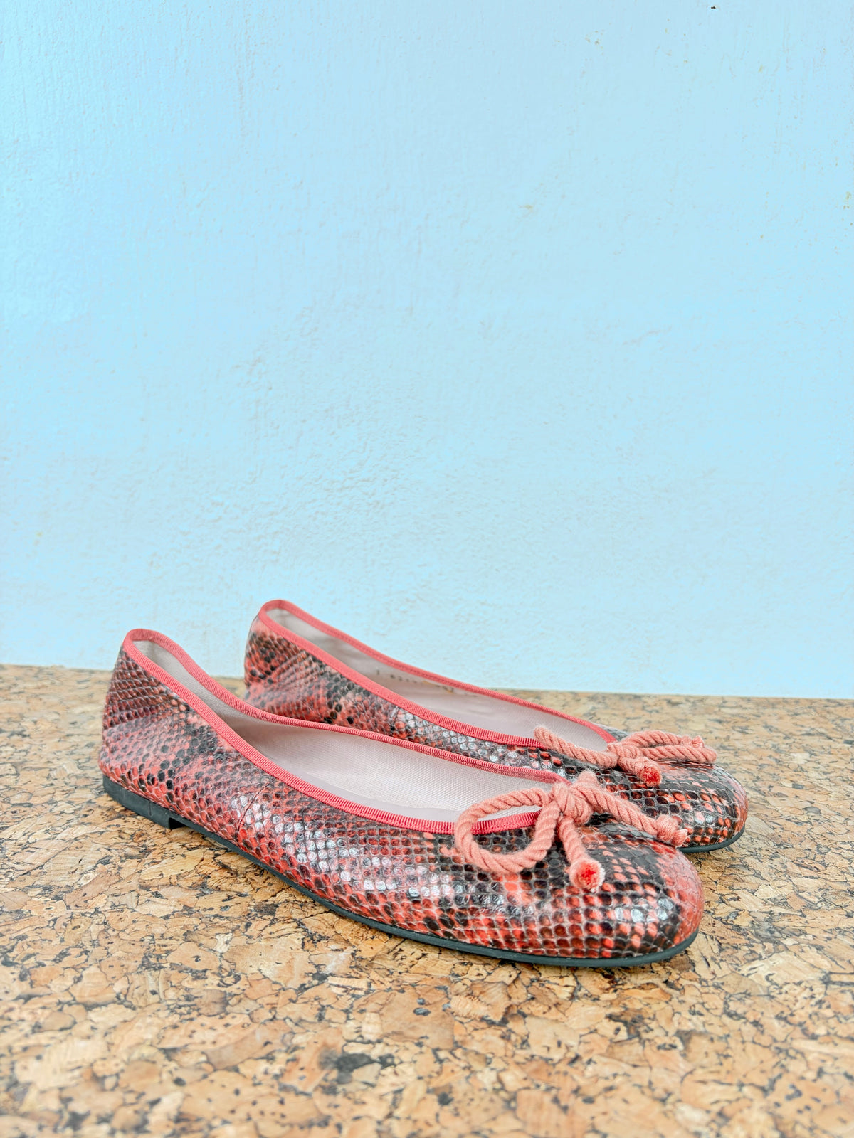 A pair of women's ballet flats with a pink and black snakeskin pattern, featuring matching pink rope bows on the toes, displayed on a cork surface against a light blue wall.