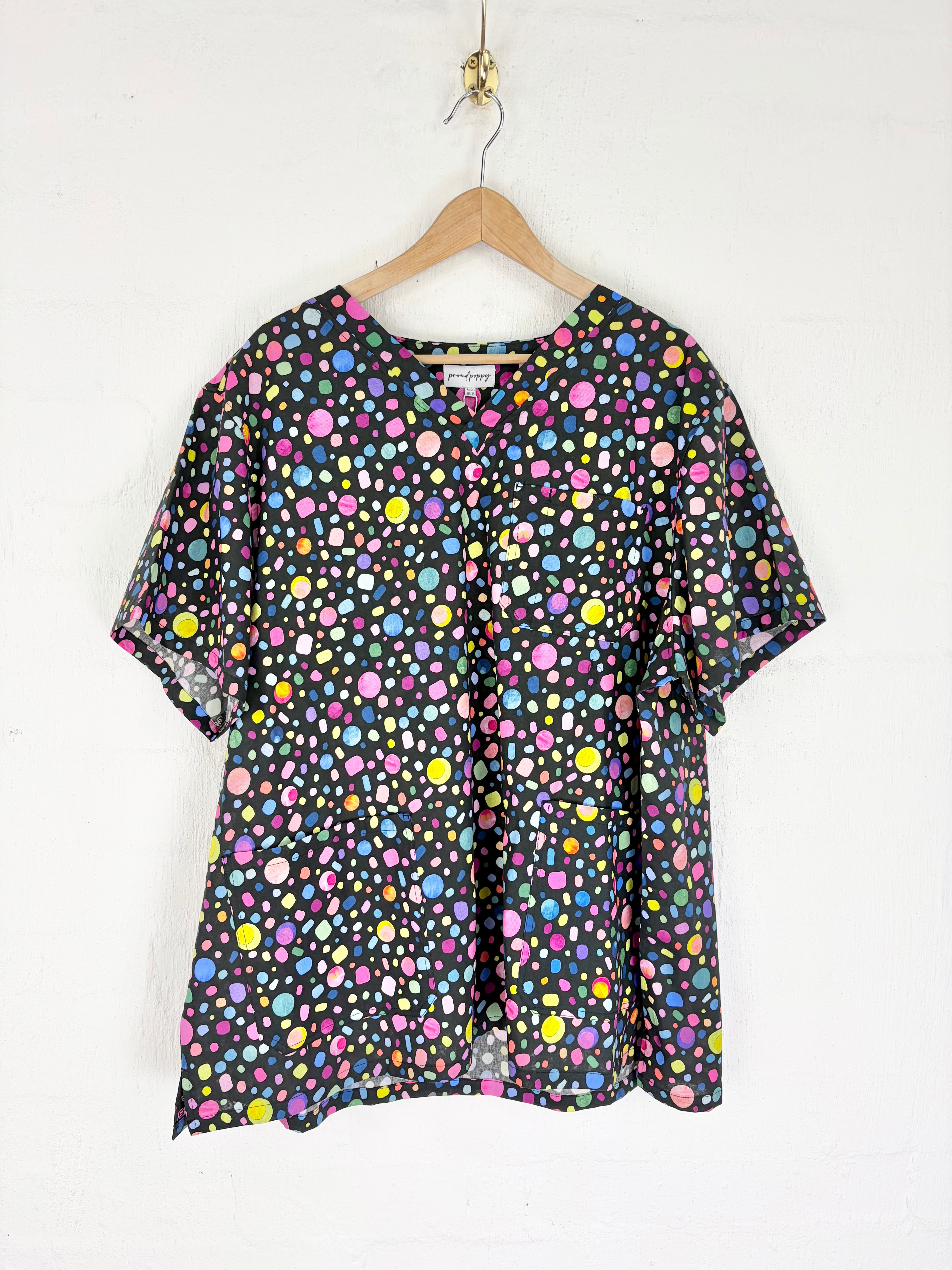 A short-sleeve button-front blouse with a colorful polka dot pattern on a black background, hanging on a wooden hanger against a white wall.