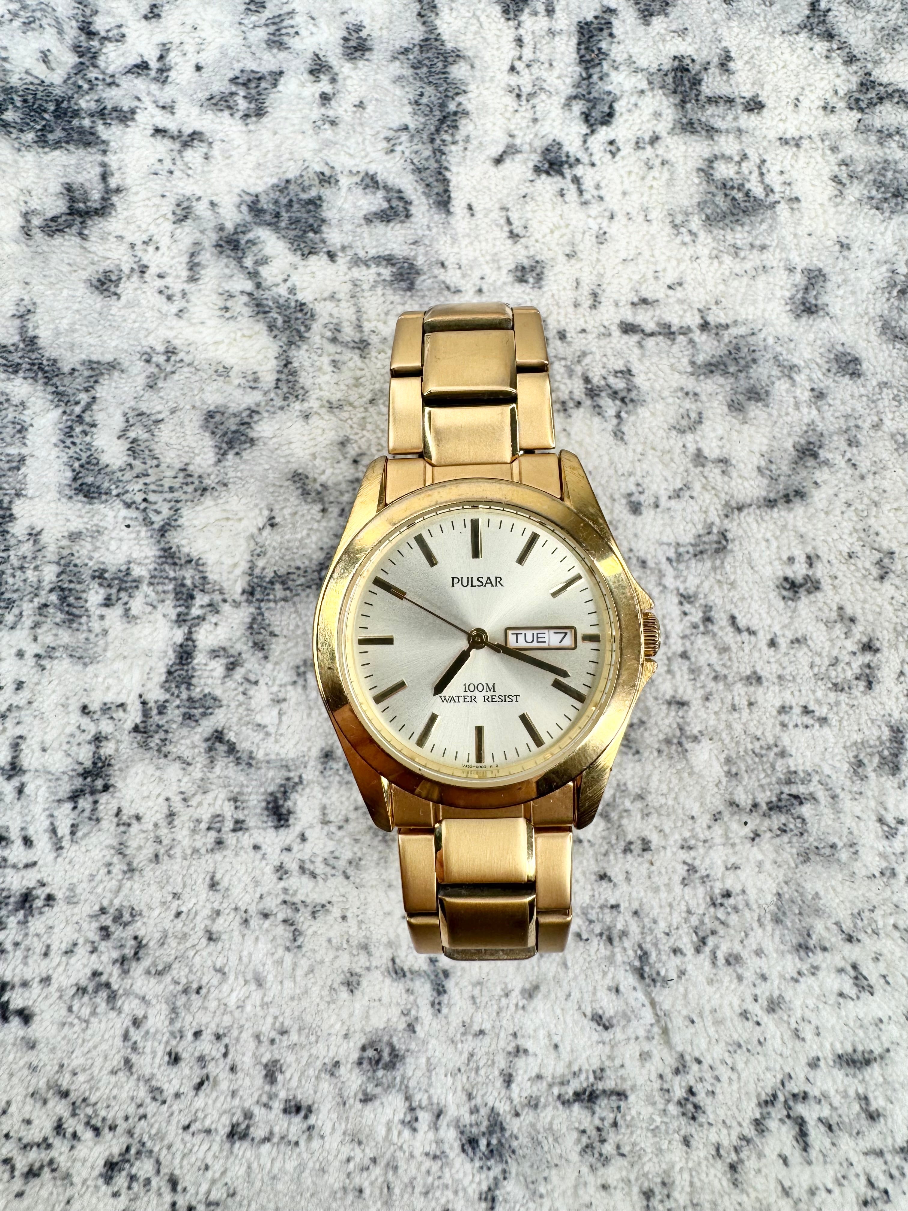 A gold-tone Pulsar wrist watch with a white dial and date window, displayed on a textured gray and white background.
