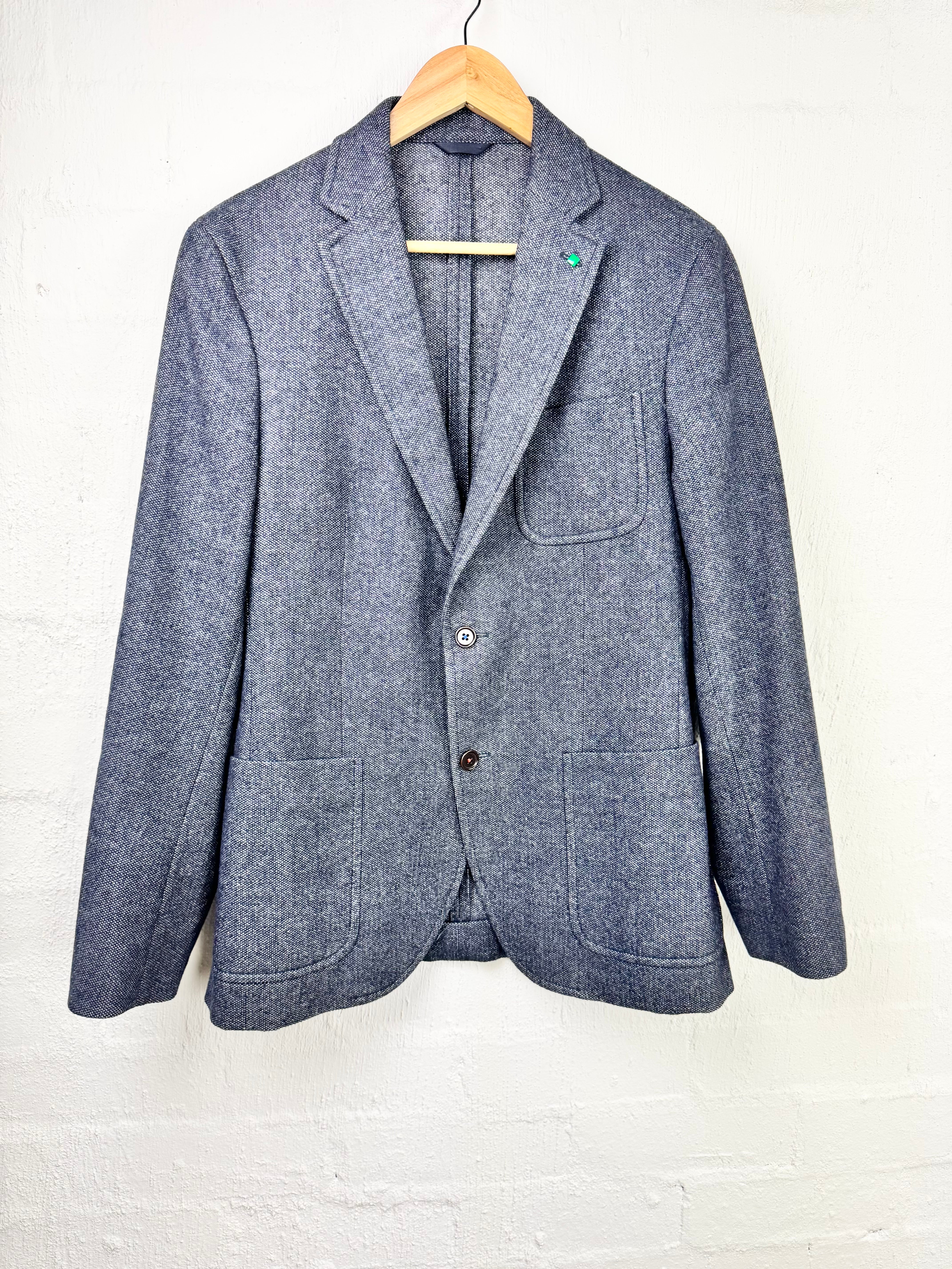 A textured blue-gray wool blazer jacket hanging on a wooden hanger against a white painted brick wall.
