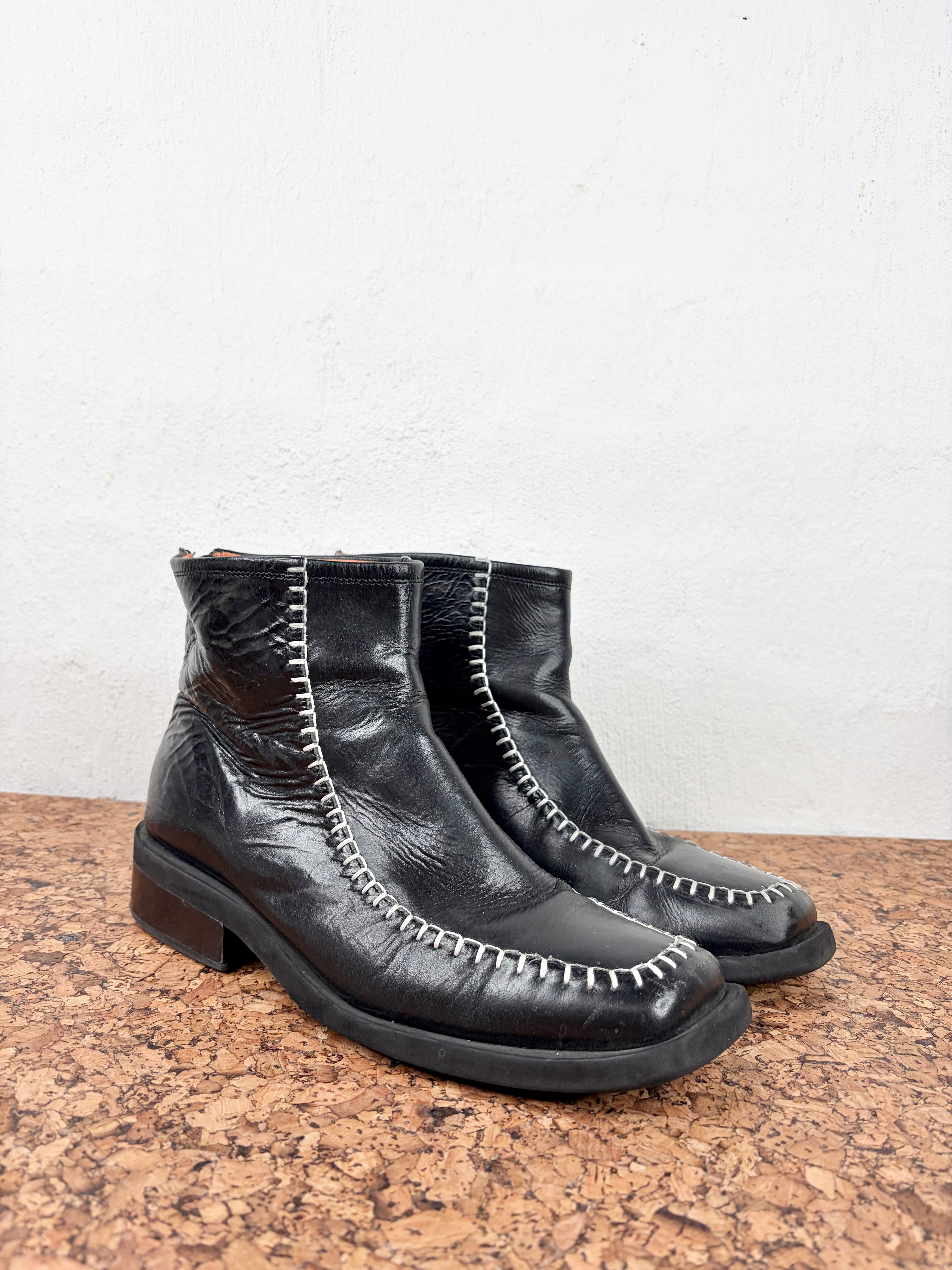 A pair of black leather ankle boots with white contrast stitching, resting on a brown cork surface against a white wall.