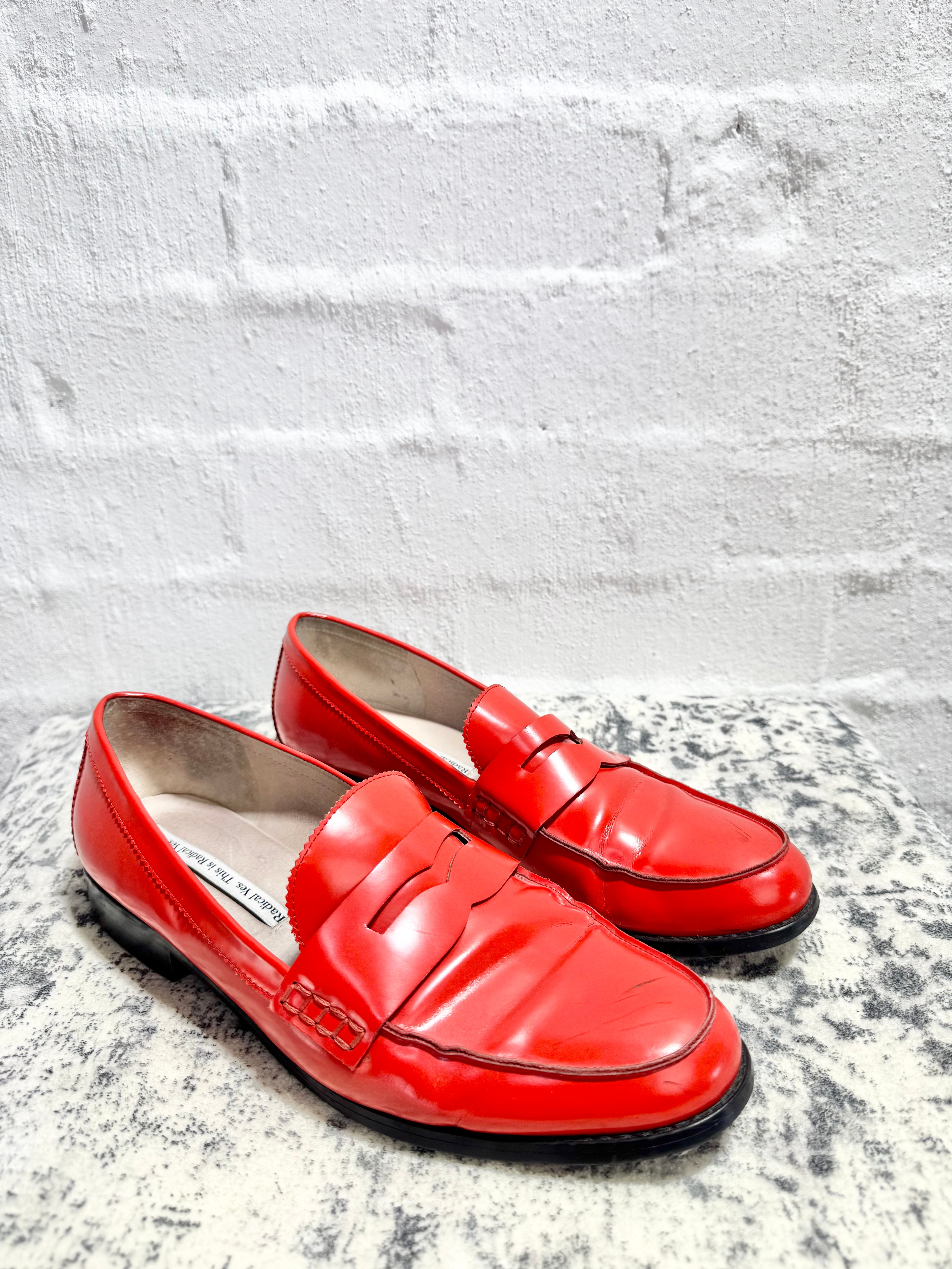 A pair of bright red leather penny loafers with black soles, displayed on a textured gray surface against a white brick wall background.