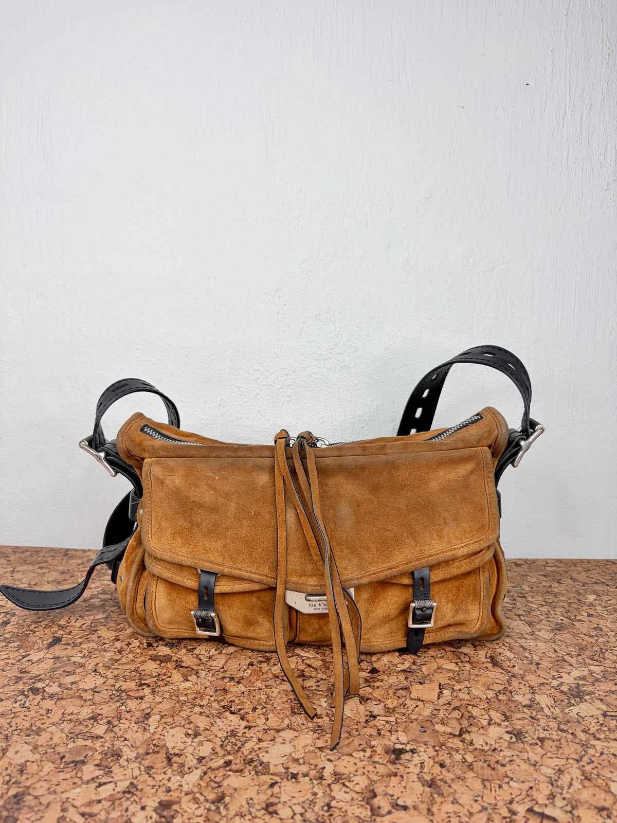 A vintage tan suede shoulder bag with black leather straps and buckles, displayed on a speckled brown countertop against a white wall.