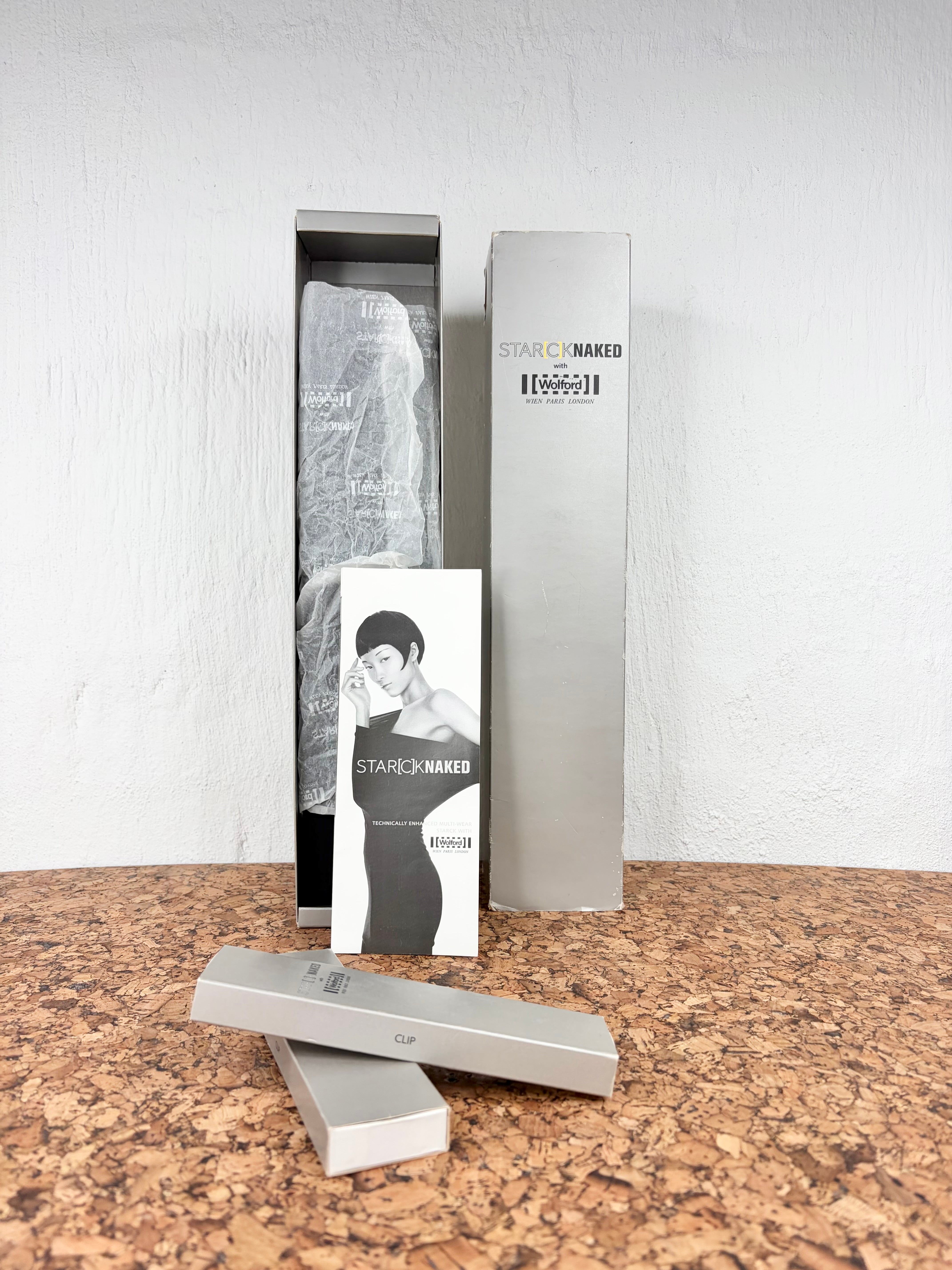 A boxed fashion item displayed on a cork-like surface against a white wall. The set includes a tall rectangular box, a slim inner package with a translucent garment, a small printed card featuring a black-and-white portrait, and two slim silver cases in front.