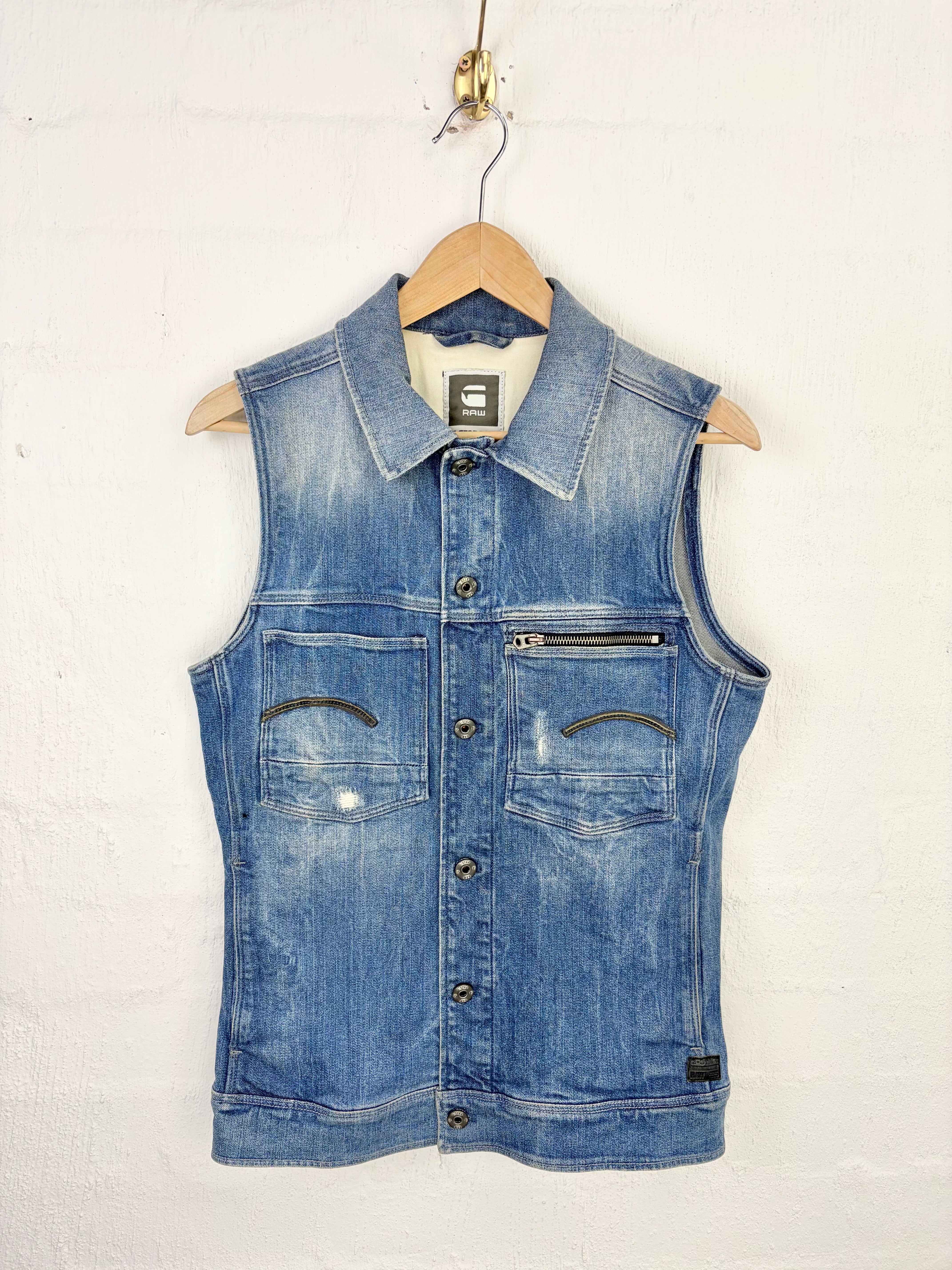 A blue denim vest with a pointed collar, button front, and zippered chest pocket, hanging on a wooden hanger against a white painted brick wall.