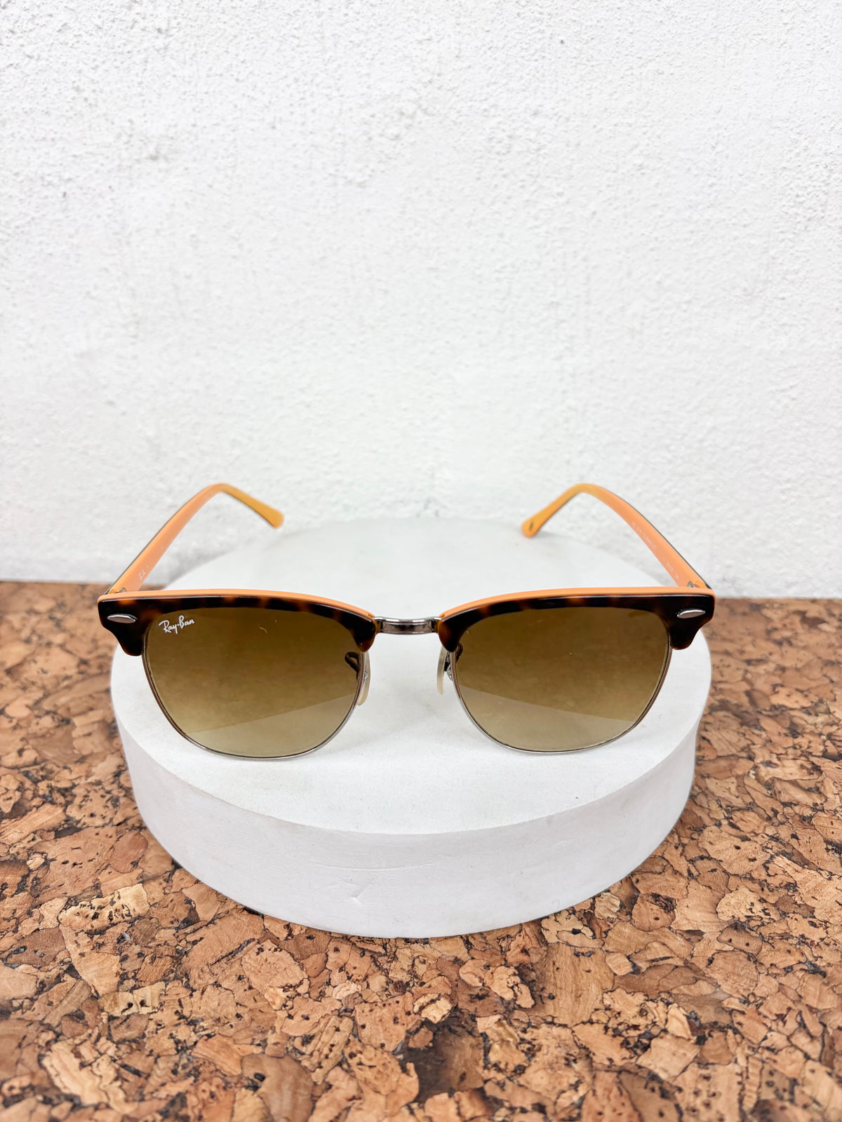 Ray-Ban Clubmaster sunglasses with a tortoiseshell pattern on the front, orange temples, and brown gradient lenses, displayed on a white cylindrical pedestal.