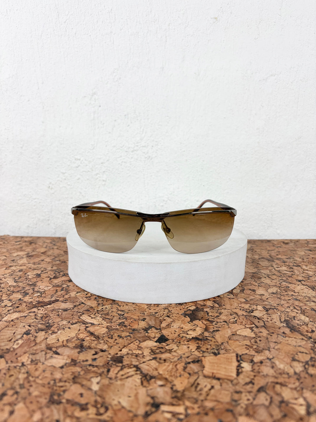 A pair of aviator-style sunglasses with a dark brown acetate frame and gradient brown lenses, displayed on a white cylindrical pedestal over a cork surface against a white wall.