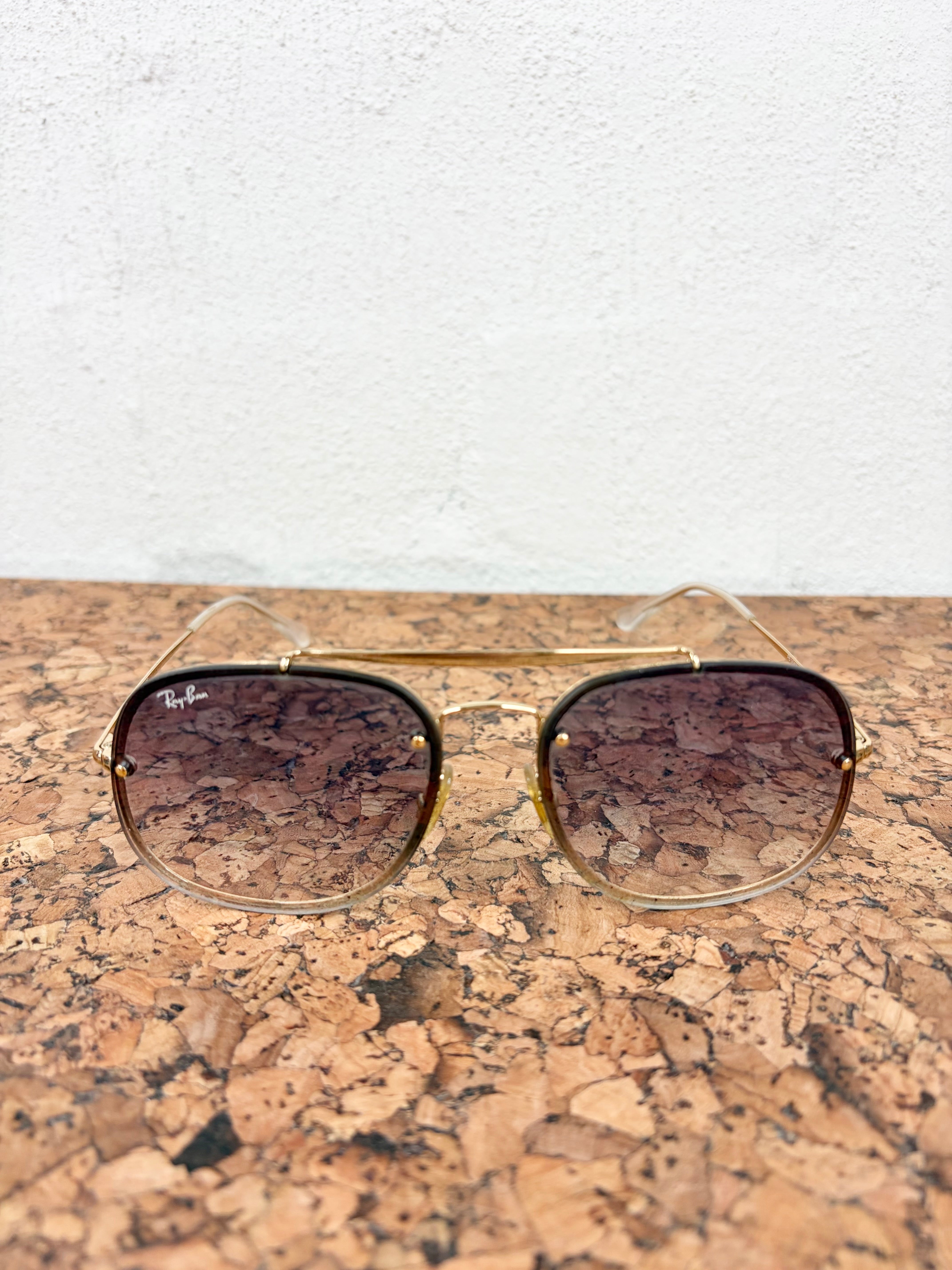Ray-Ban aviator sunglasses with gold metal frames and gradient brown lenses, resting on a cork surface against a white wall.
