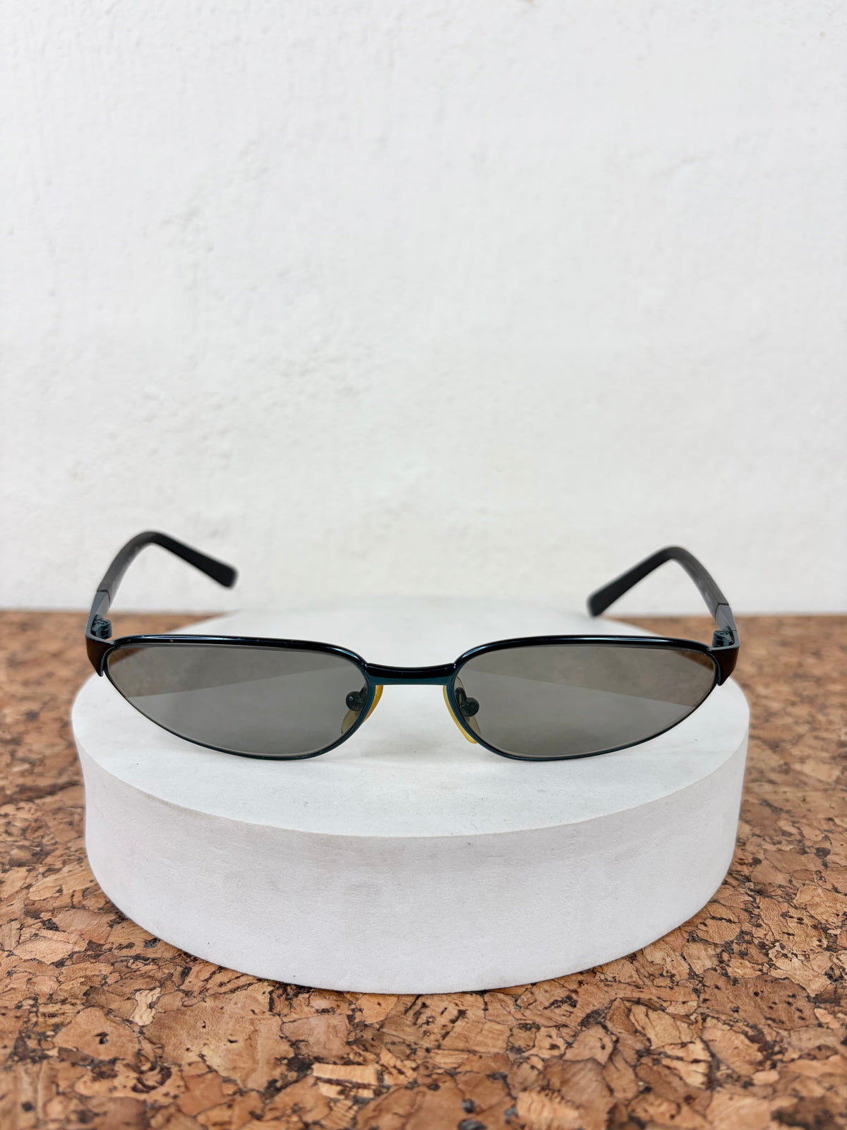 Oval sunglasses with a black frame and dark gray lenses, displayed on a round white pedestal over a cork surface against a white wall.