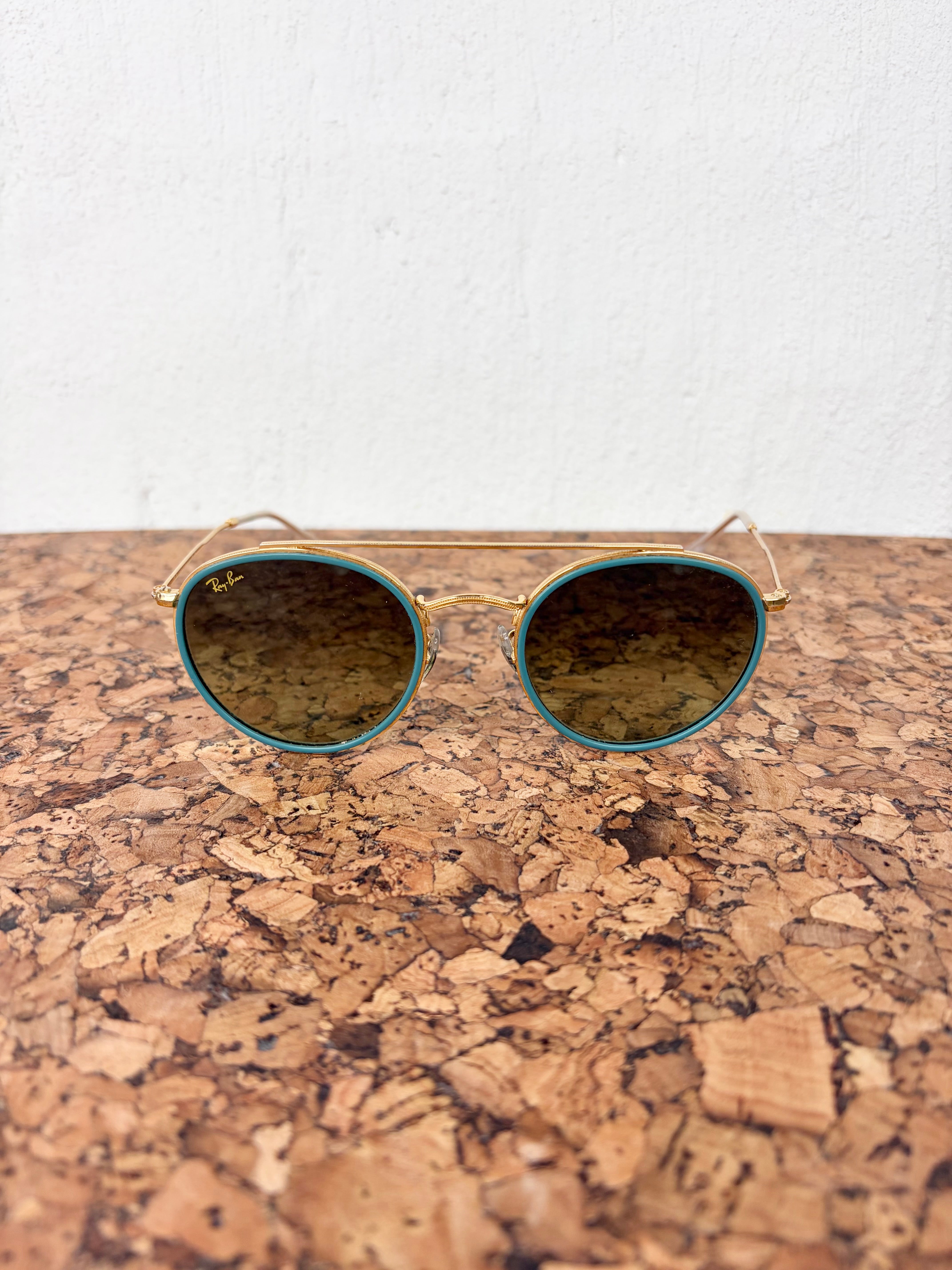 Round aviator-style sunglasses with gold metal frames and dark brown tinted lenses, resting on a cork surface against a white wall.