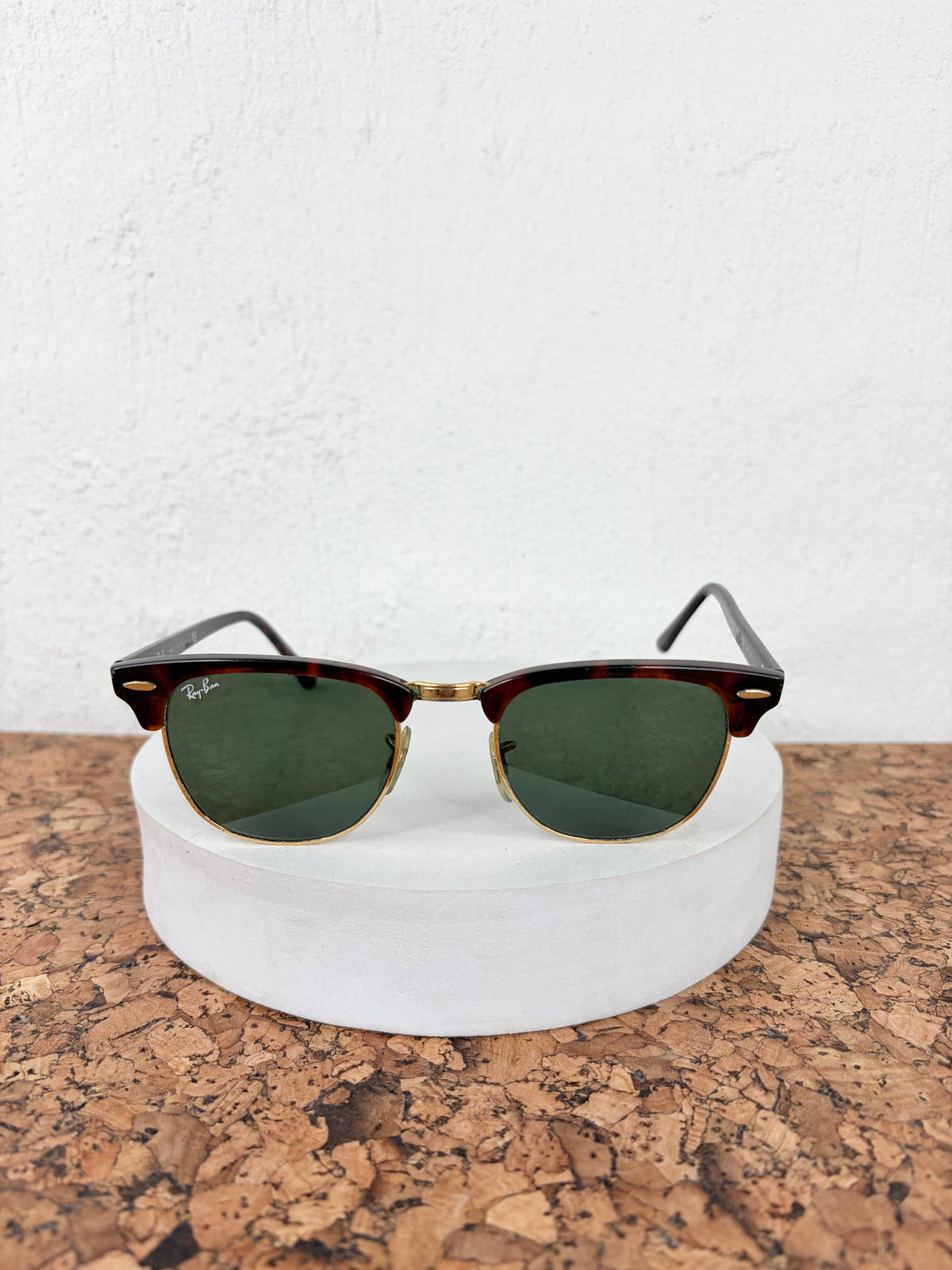 Ray-Ban Clubmaster sunglasses with dark green lenses, brown acetate frames, and gold metal rims, displayed on a white cylindrical stand over a cork surface against a white wall.