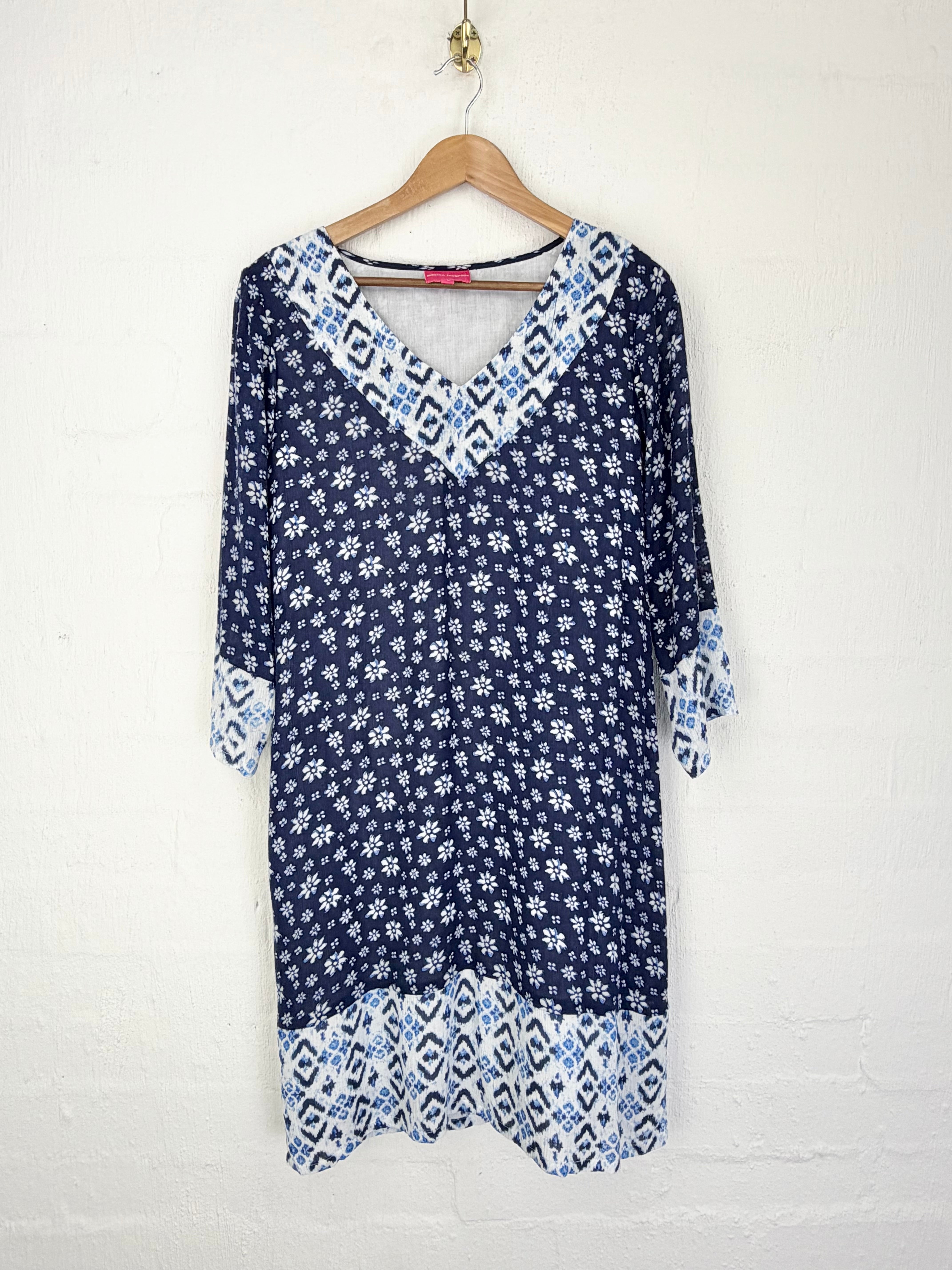 A navy blue and white patterned tunic top with geometric and floral prints, hanging on a wooden hanger against a white painted brick wall.