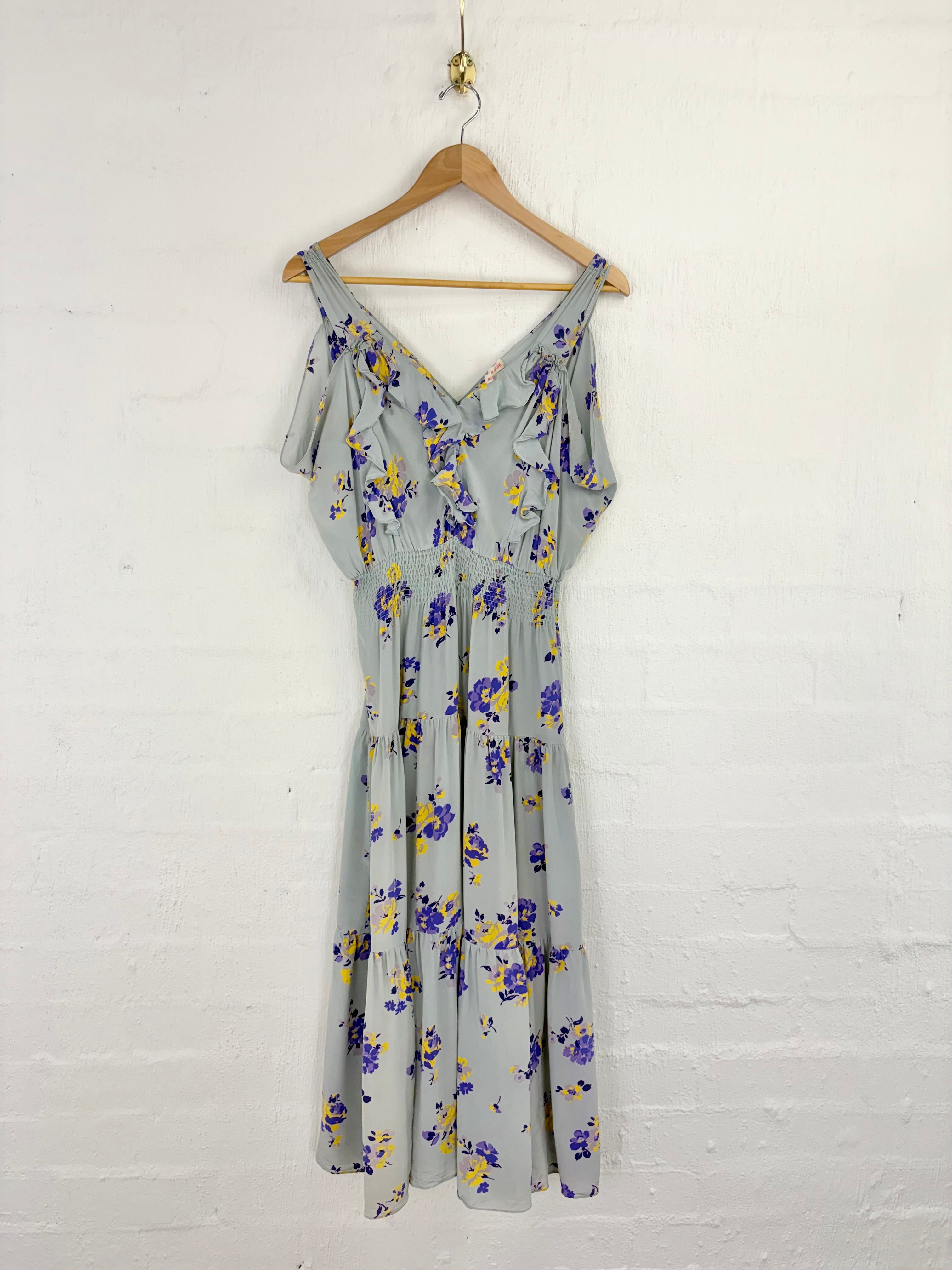 A light gray sleeveless dress with a purple and yellow floral print, hanging on a wooden hanger against a white painted brick wall.