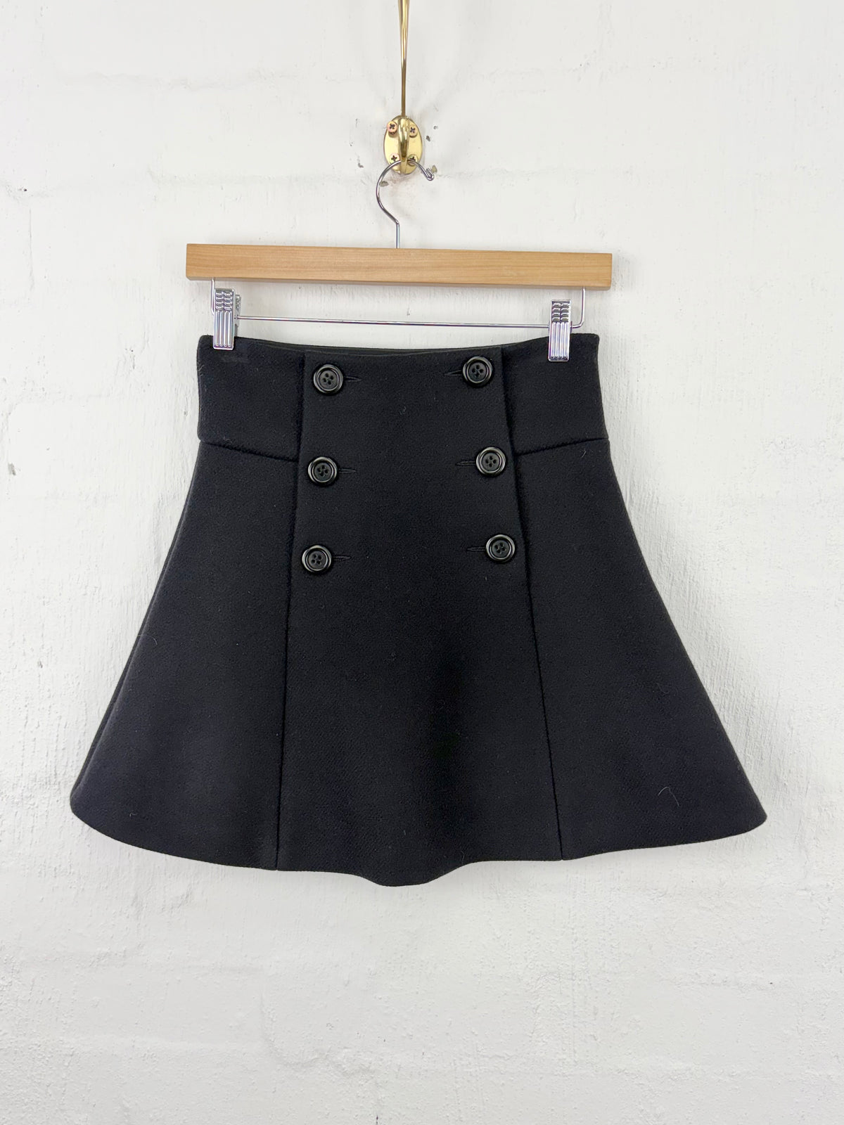 A black high-waisted flared mini skirt with double-breasted buttons, hanging on a wooden hanger against a white painted brick wall.