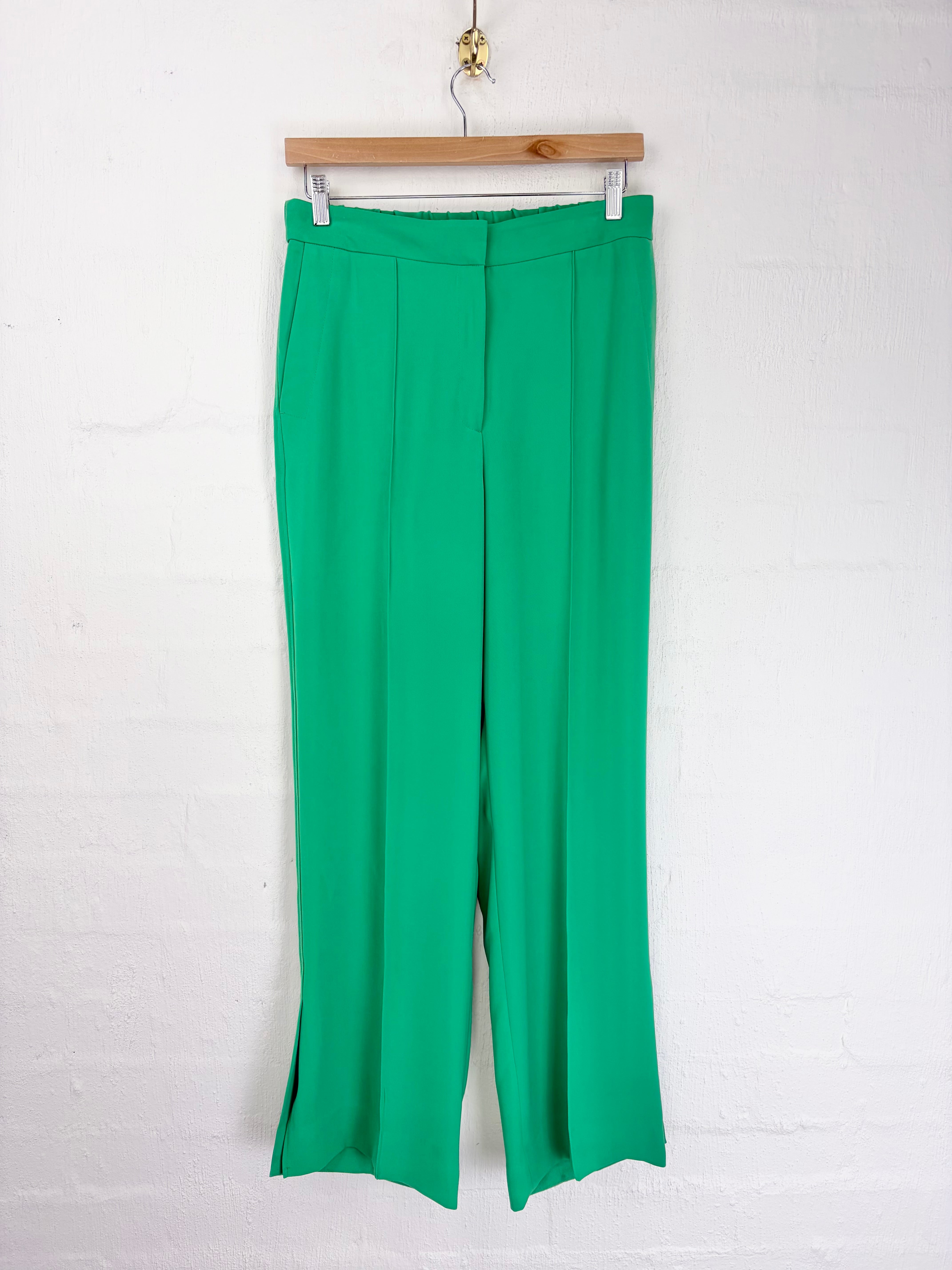 Green wide-leg cropped trousers hanging from a wooden hanger against a white brick wall.