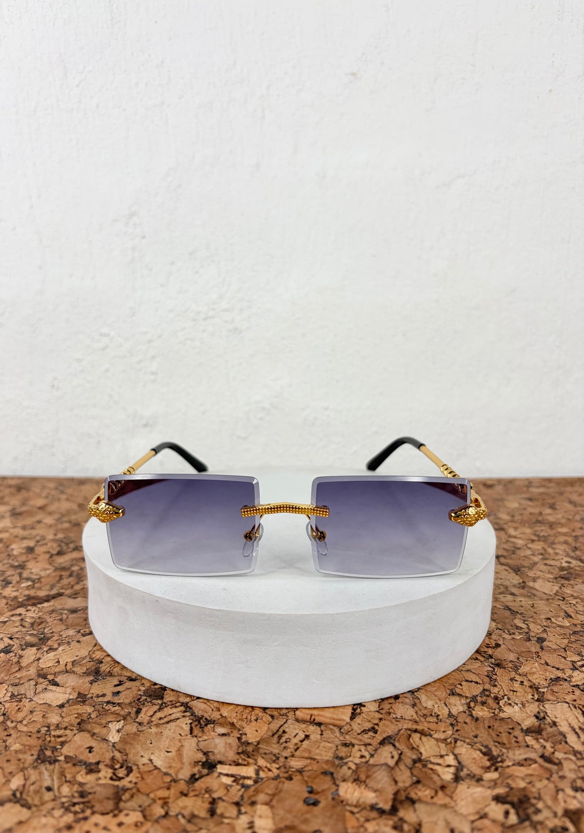 Rimless square sunglasses with gold metal frames and gradient purple lenses, displayed on a round white pedestal over a cork surface against a white wall.