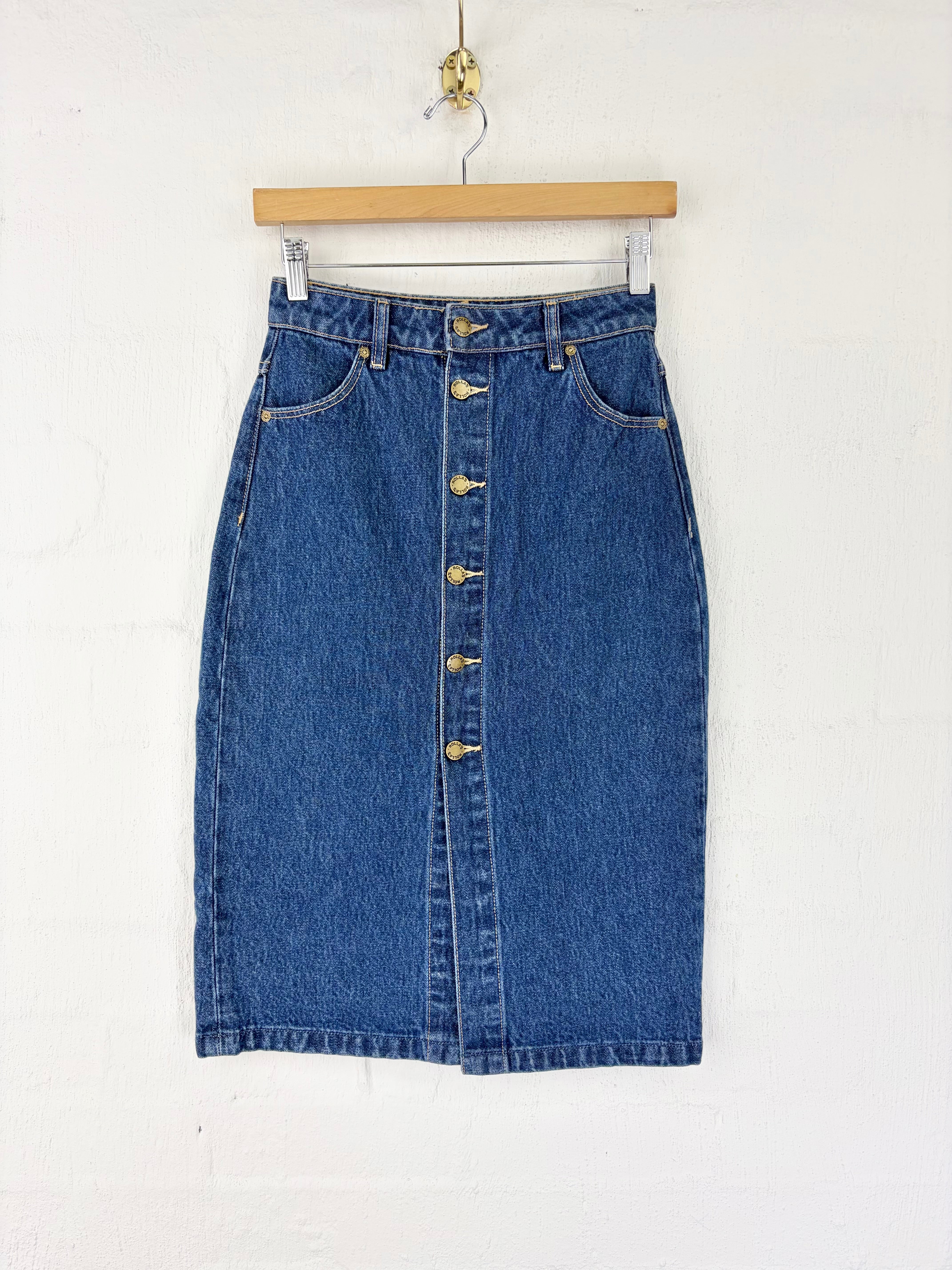 A blue denim pencil skirt with a front button closure, hanging from a wooden hanger against a white wall.