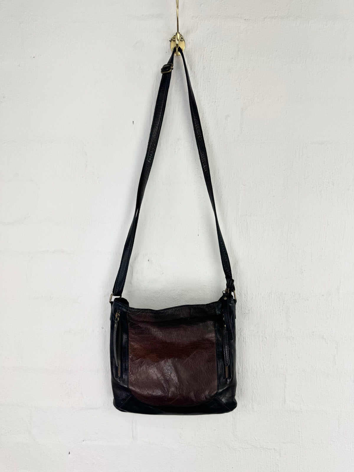 A dark brown and black leather shoulder bag hanging from a gold hook against a white painted brick wall.
