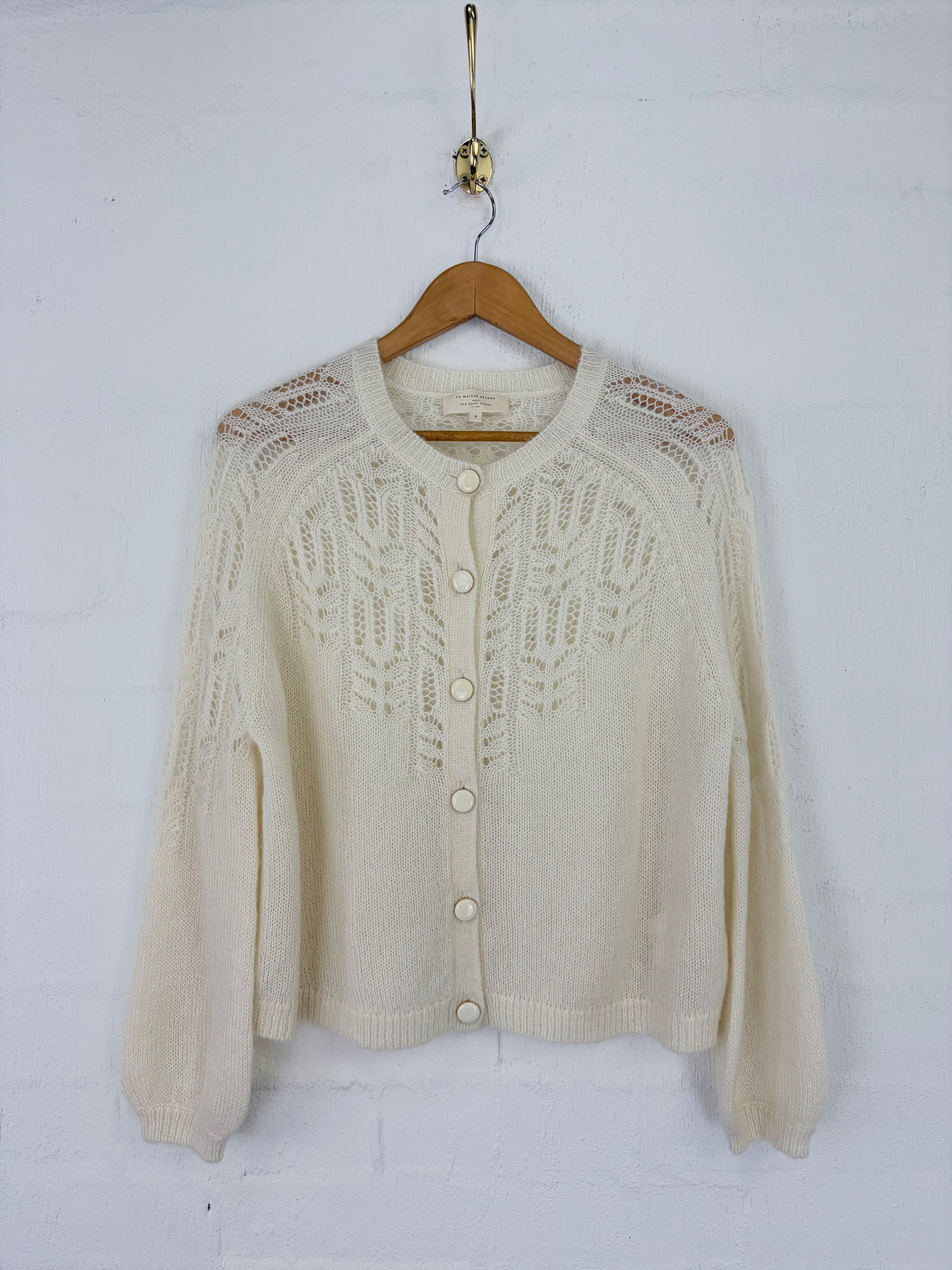 An off-white/cream cable-knit cardigan sweater with button-front closure and long sleeves, hanging on a wooden hanger against a white painted brick wall.