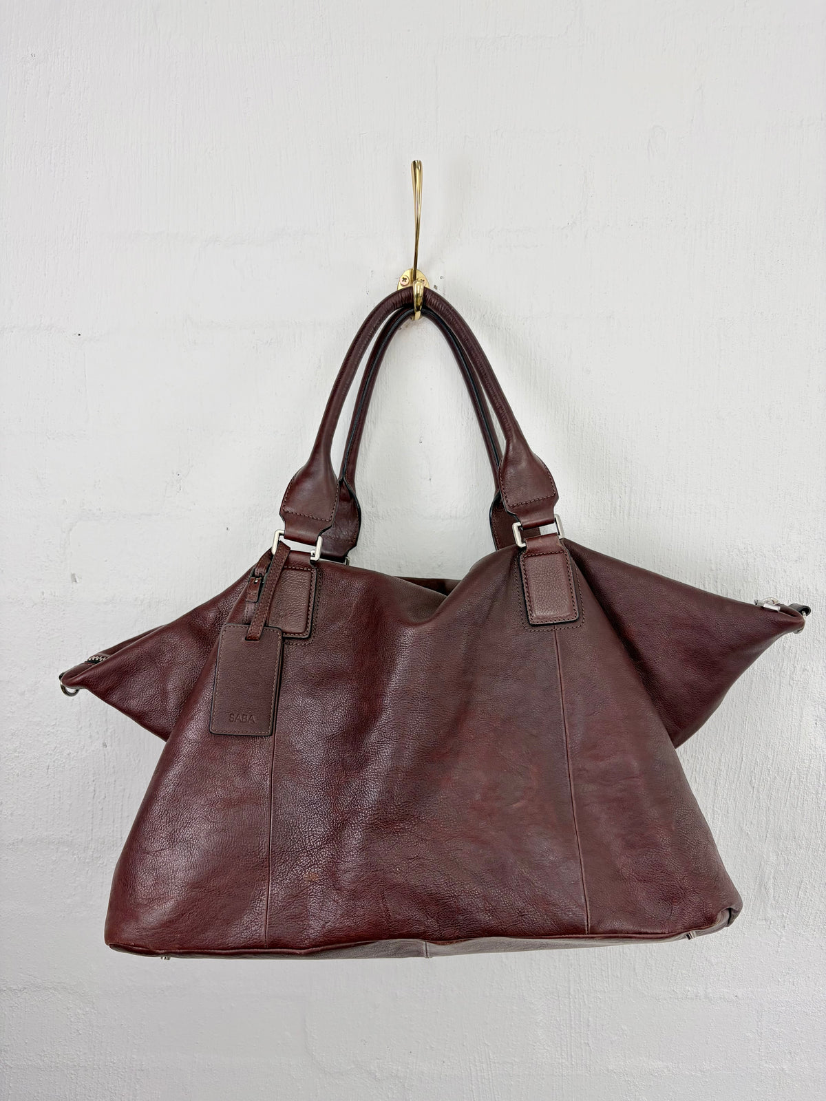 A dark reddish-brown leather tote bag with double top handles and silver-tone hardware, hanging from a gold hook against a white textured wall.