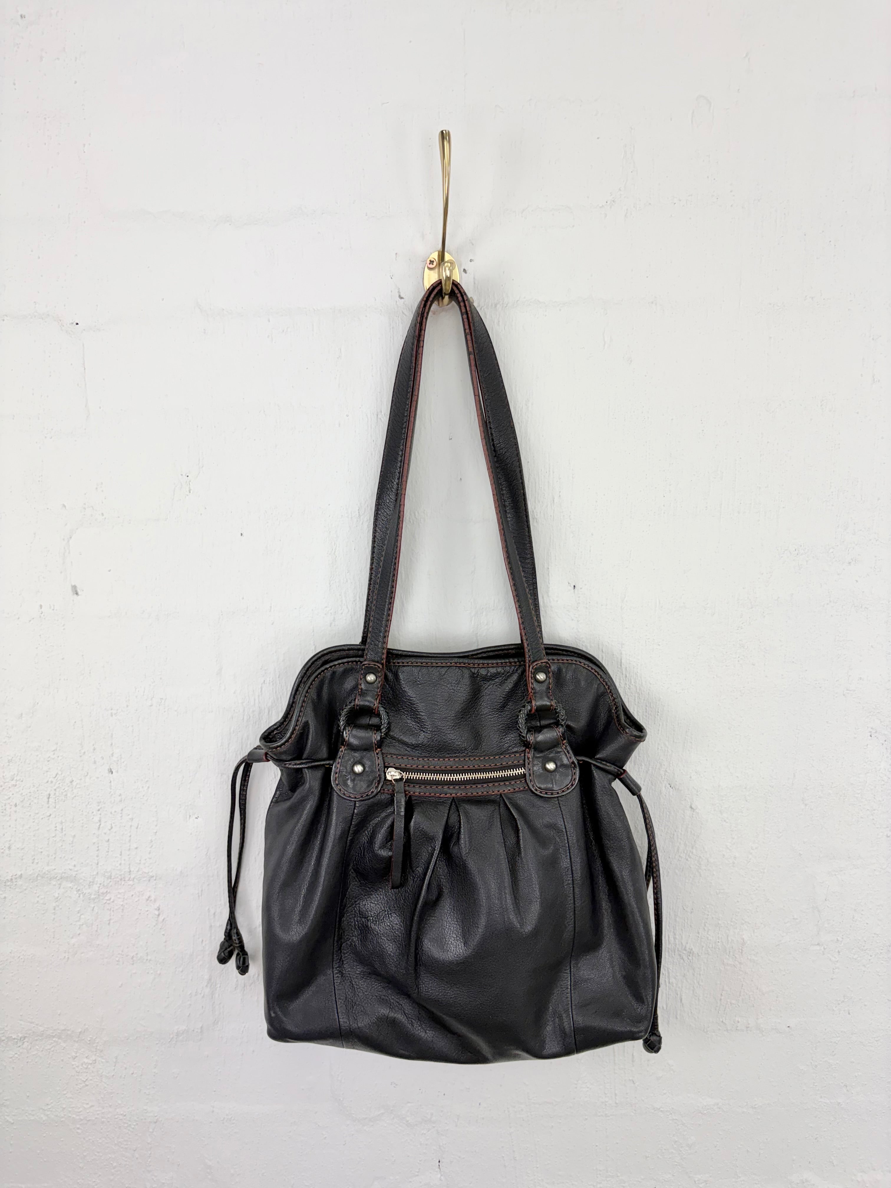 Black leather shoulder bag with gold-tone hardware, hanging from a gold hook against a white painted brick wall.