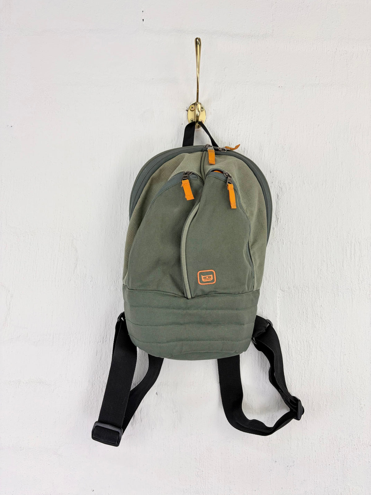 An olive green backpack with black straps and bright orange zipper pulls, hanging from a gold hook against a white wall.