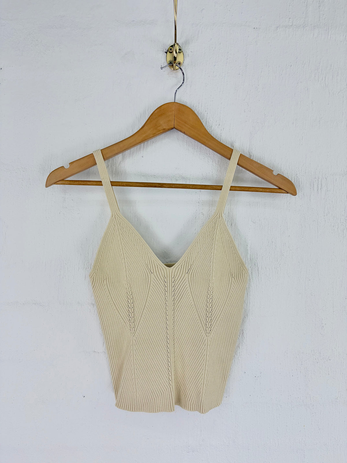 A beige ribbed knit camisole top with thin straps and a V-neckline, hanging on a wooden hanger against a white textured wall.