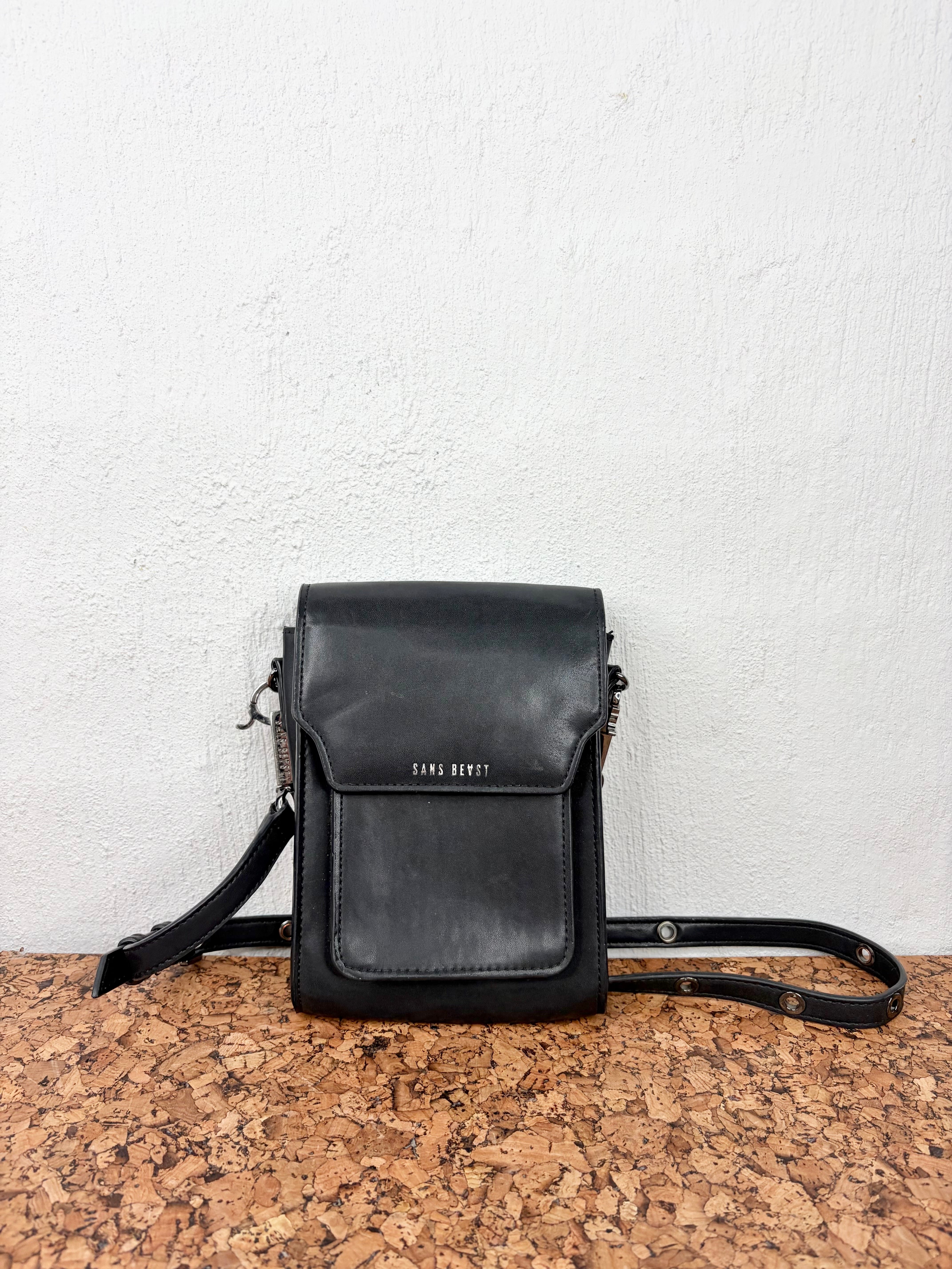 A black leather crossbody bag with a front flap pocket and silver-tone hardware, displayed on a cork surface against a white textured wall. The brand name 'TARO DOKKI' is printed in silver on the front pocket.