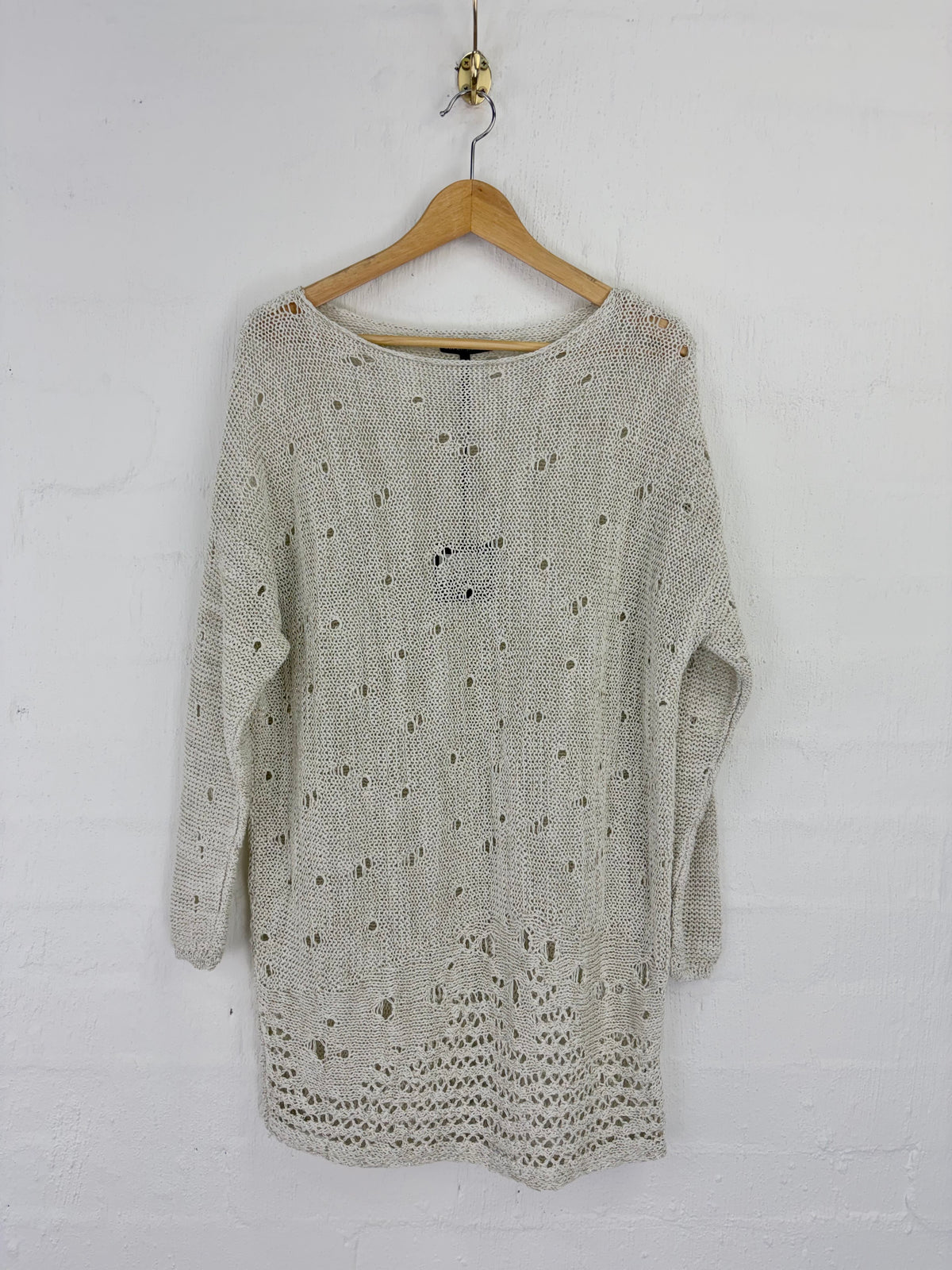A light beige knit sweater with a textured openwork pattern, hanging on a wooden hanger against a white painted brick wall.