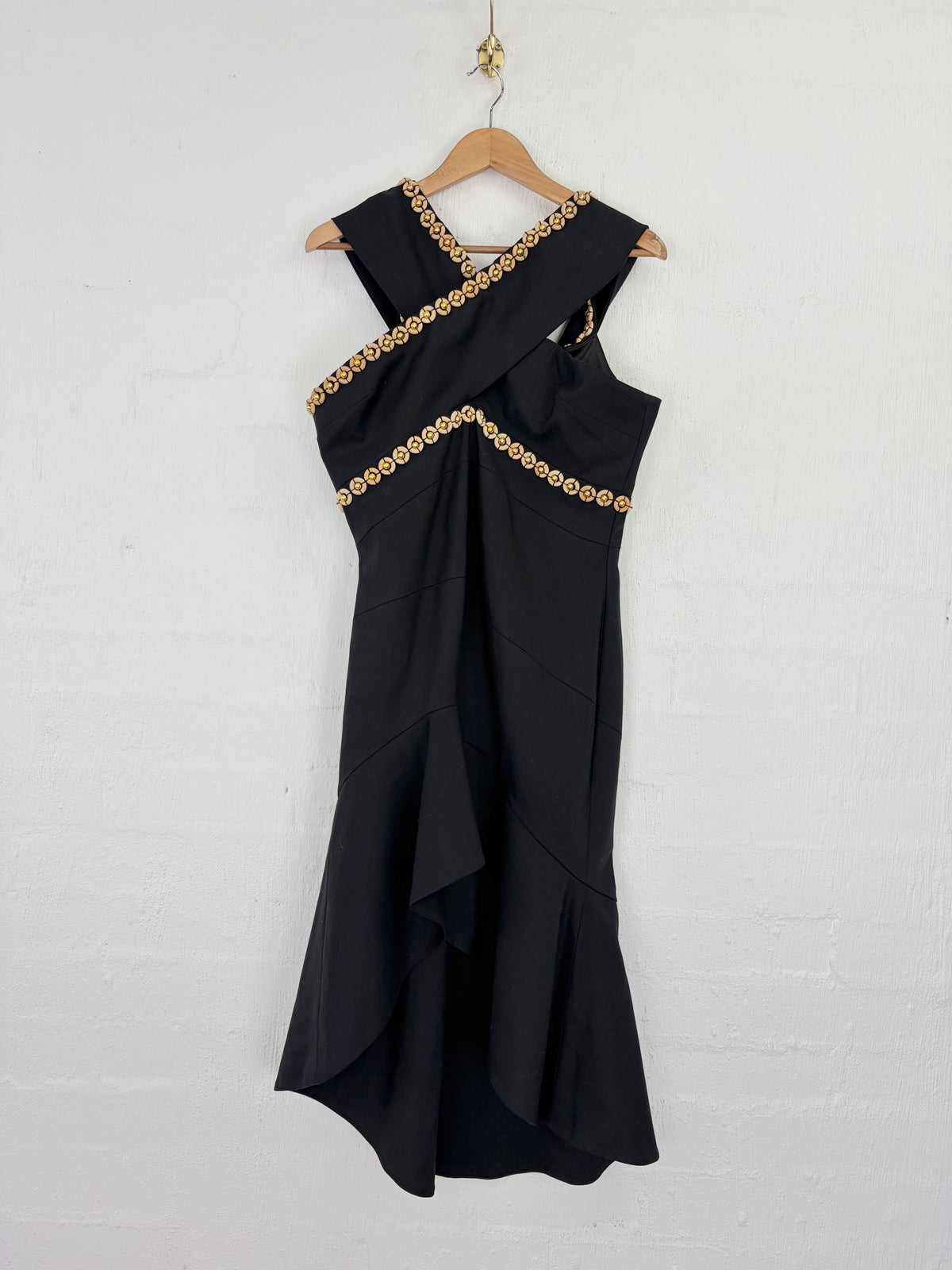 A sleeveless black dress with gold circular embellishments along the neckline and waist, hanging on a wooden hanger against a white brick wall.