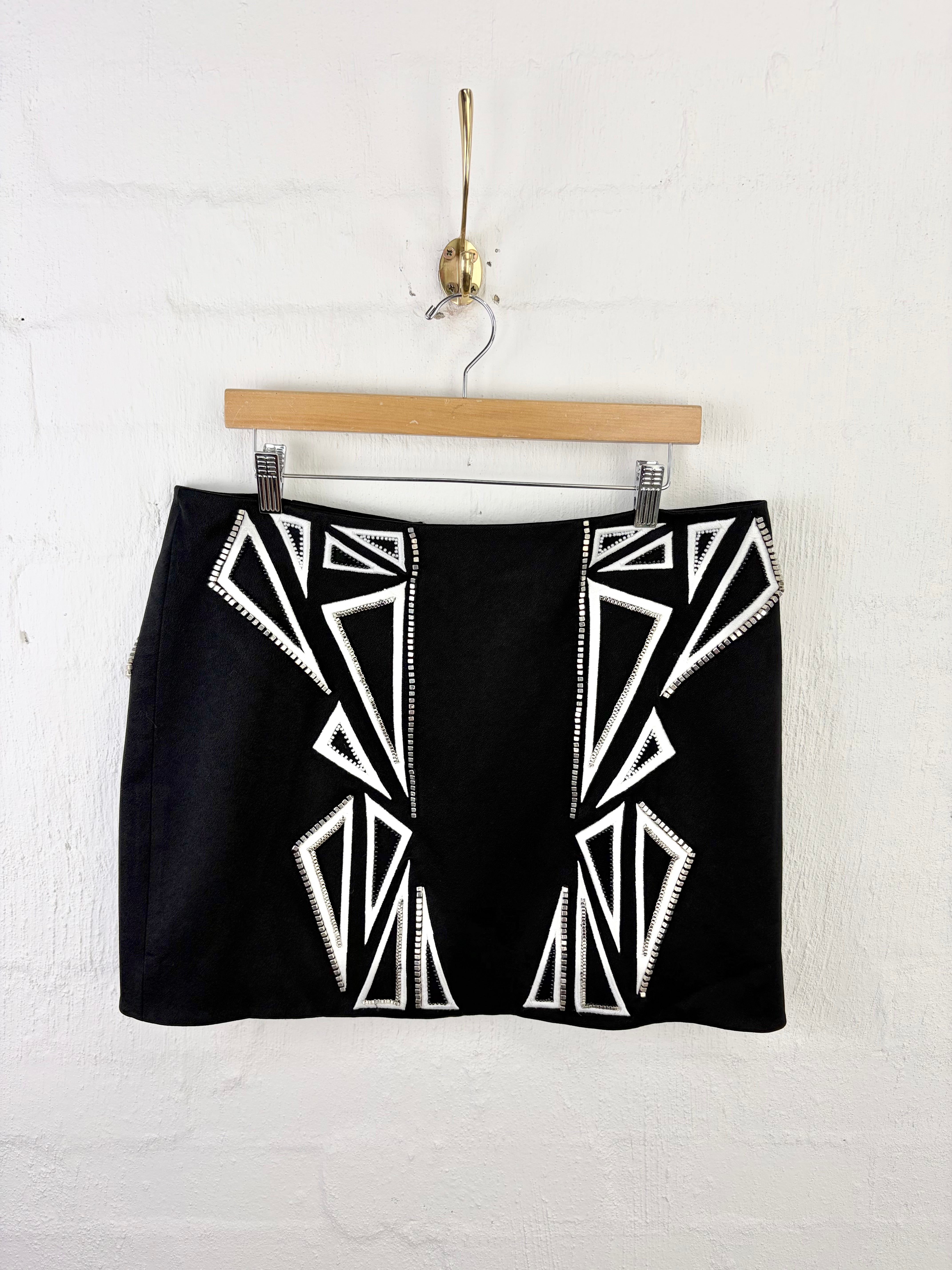 A black mini skirt with white geometric Art Deco style embroidery, hanging from a wooden hanger against a white painted brick wall.