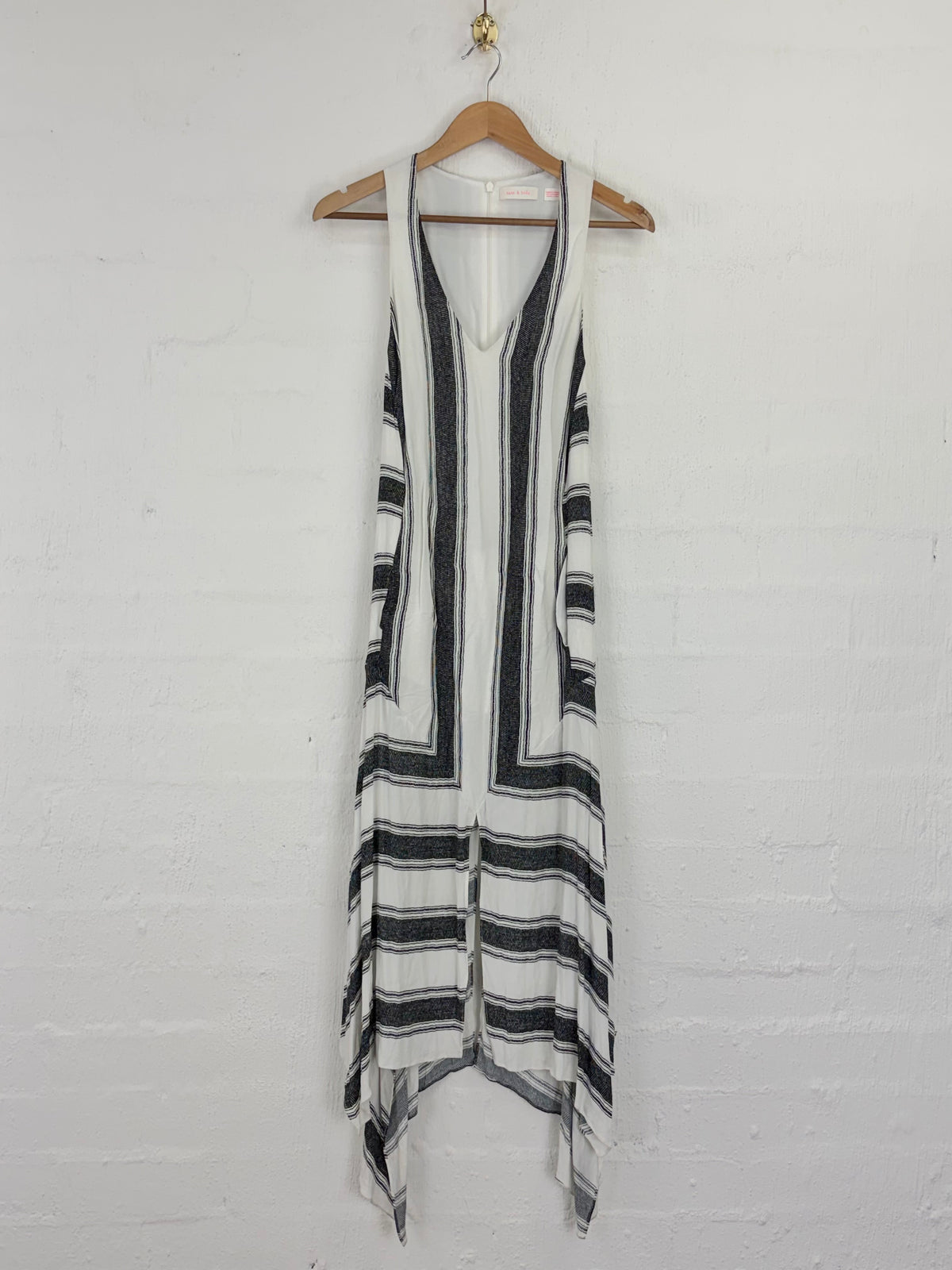 A sleeveless knit dress with black and white vertical stripes and horizontal bands near the hem, hanging on a wooden hanger against a white painted brick wall.