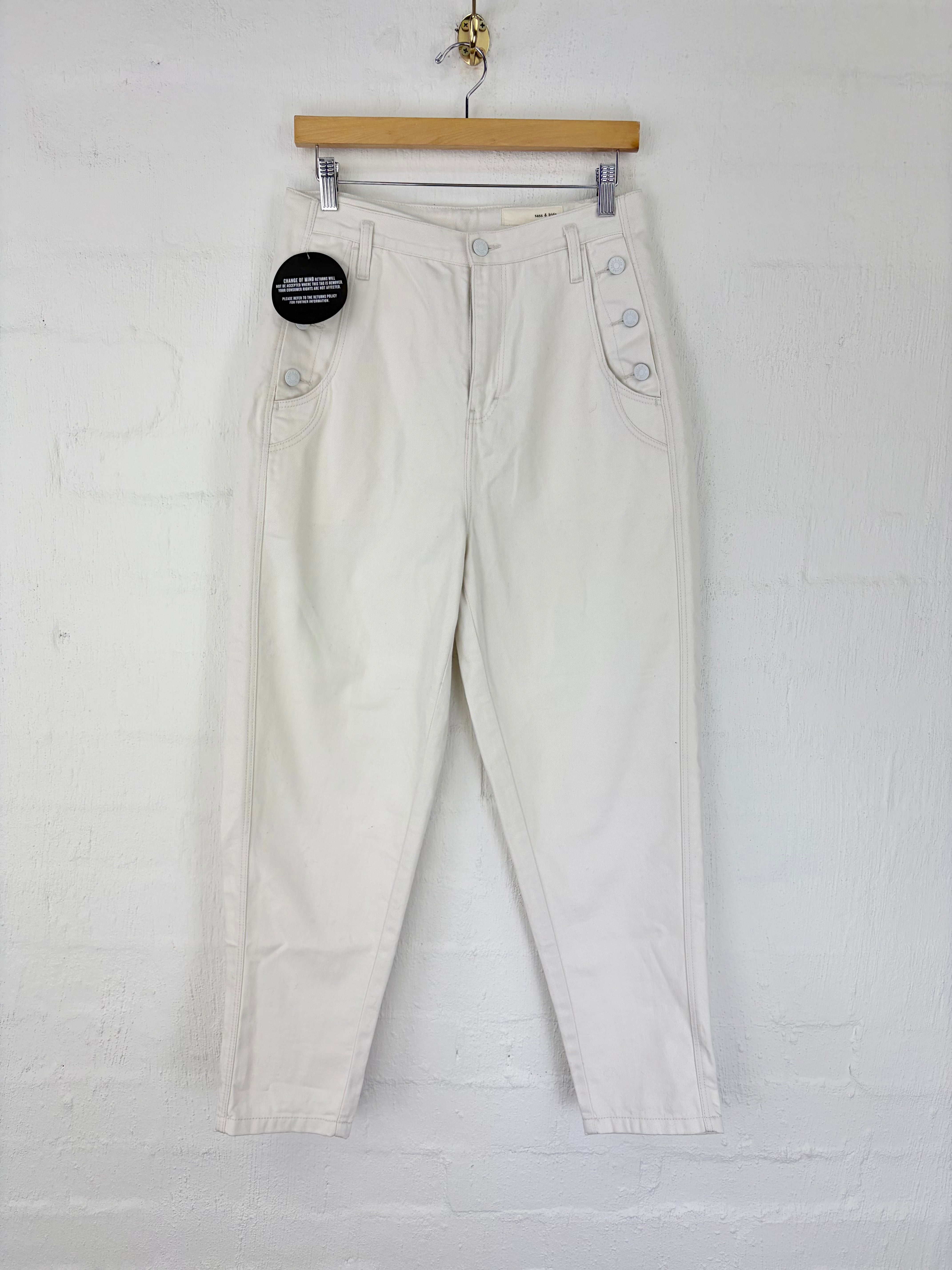 Off-white cropped pants with decorative side buttons, hanging from a wooden hanger against a white brick wall.