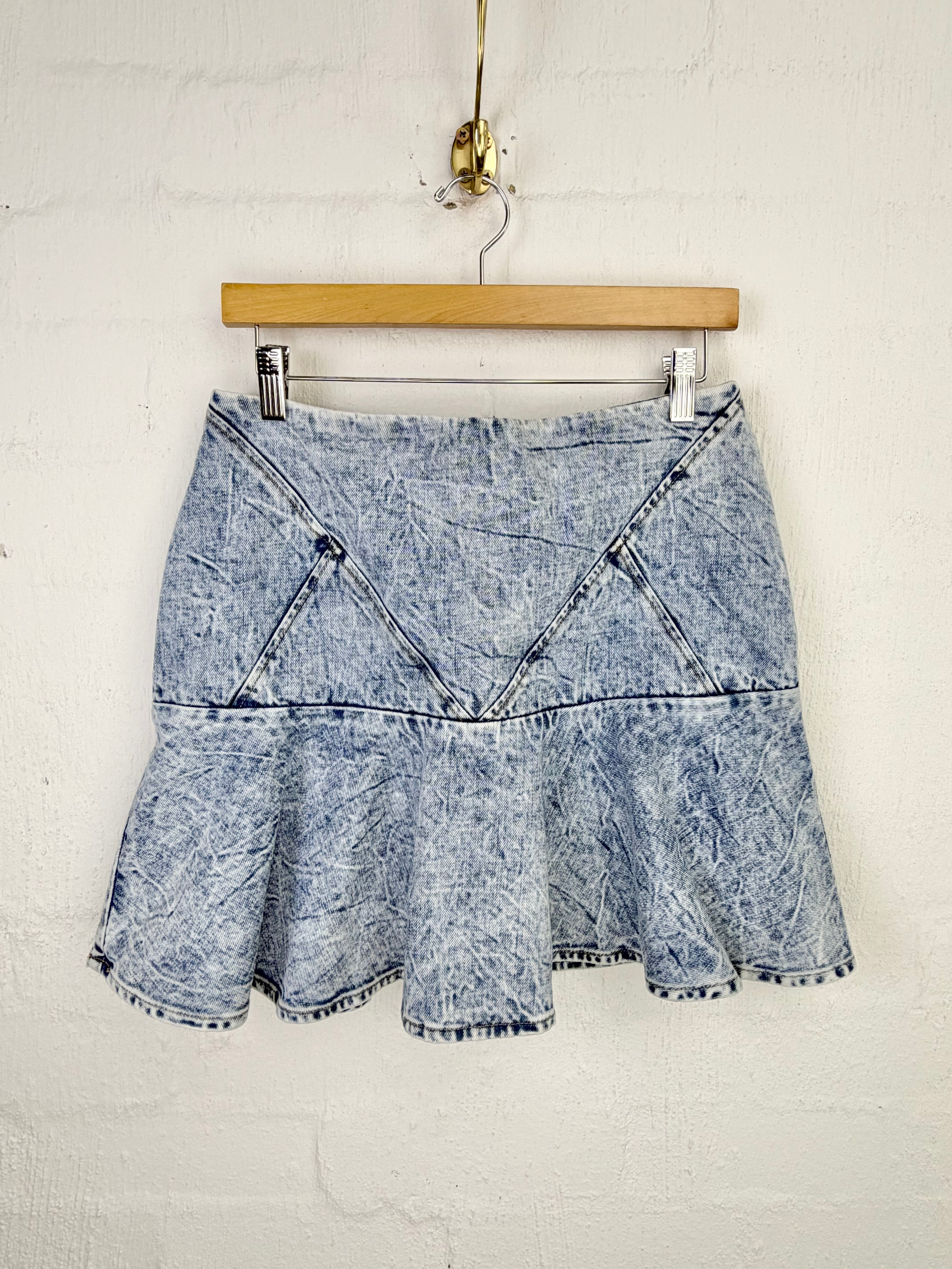 A light blue acid-wash denim mini skirt with a ruffled hem and diagonal seam details, hanging from a wooden hanger against a white painted brick wall.
