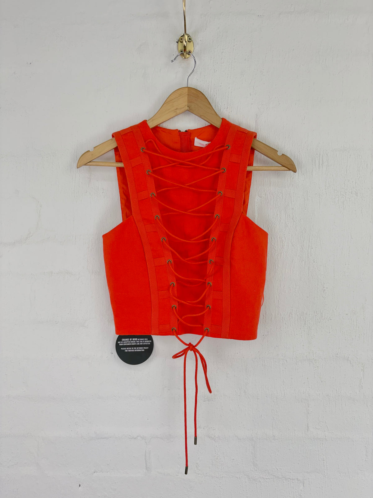 A bright orange-red sleeveless lace-up vest hanging on a wooden hanger against a white painted brick wall. The vest has a structured corset-style back with multiple eyelets and red laces, and a small black circular hangtag is attached near the lower left side.