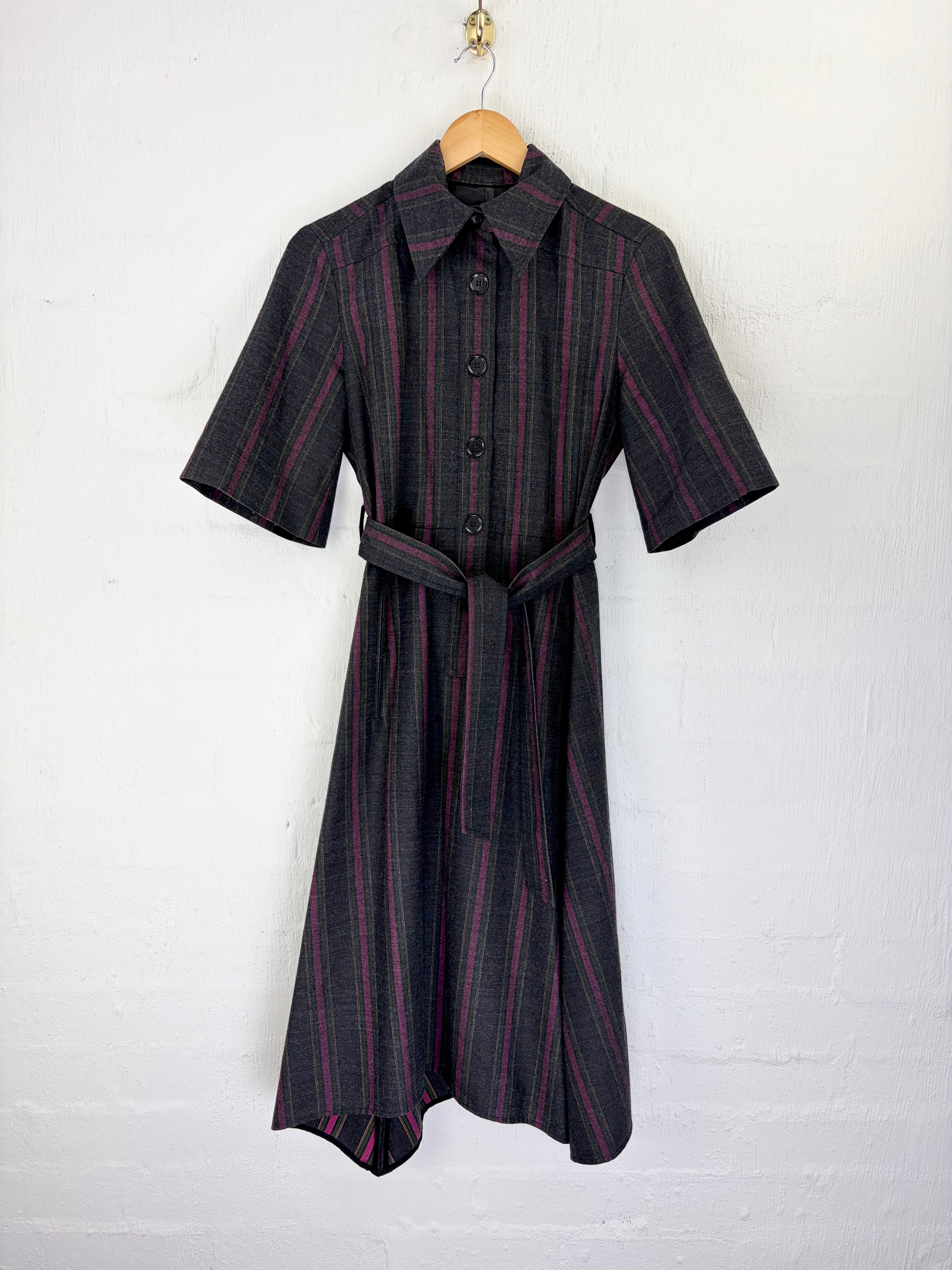 A short-sleeve collared shirt dress with a matching waist tie, featuring vertical dark pink stripes on a black and gray plaid fabric, hanging on a wooden hanger against a white painted brick wall.