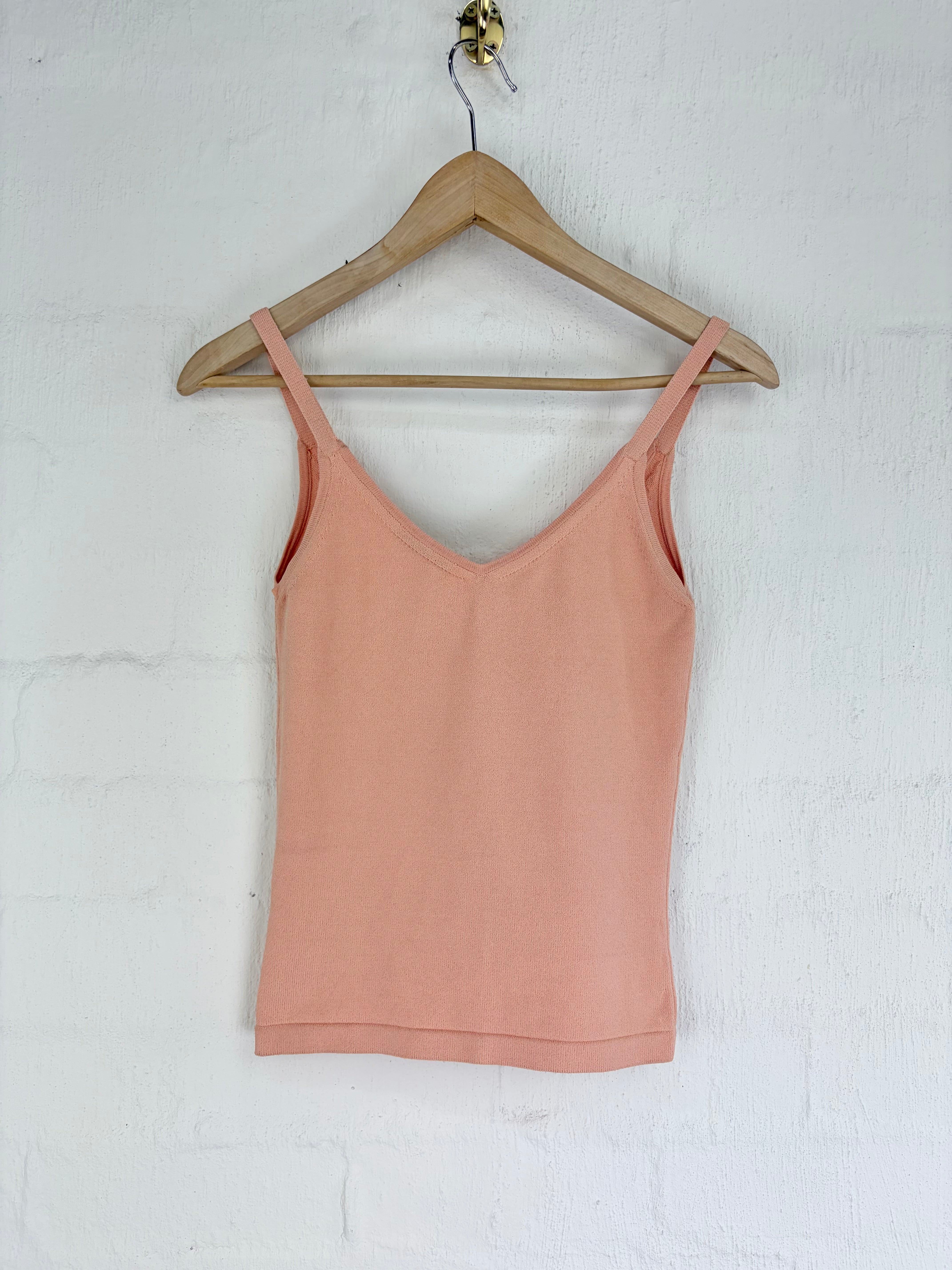 A peach-colored spaghetti strap tank top hanging on a wooden hanger against a white painted brick wall.
