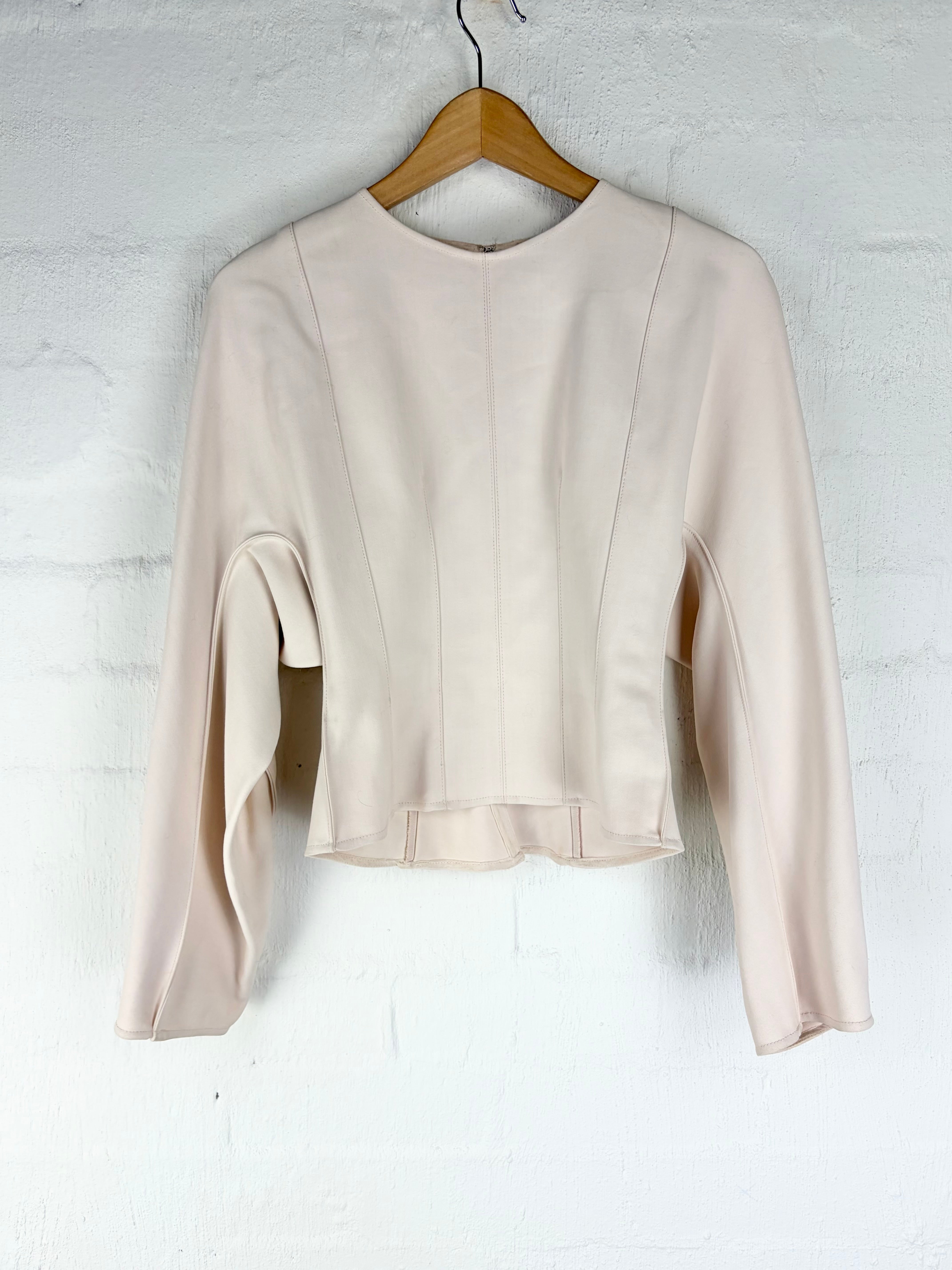 A light beige long-sleeve cropped top with a round neckline, hanging on a wooden hanger against a white brick wall.
